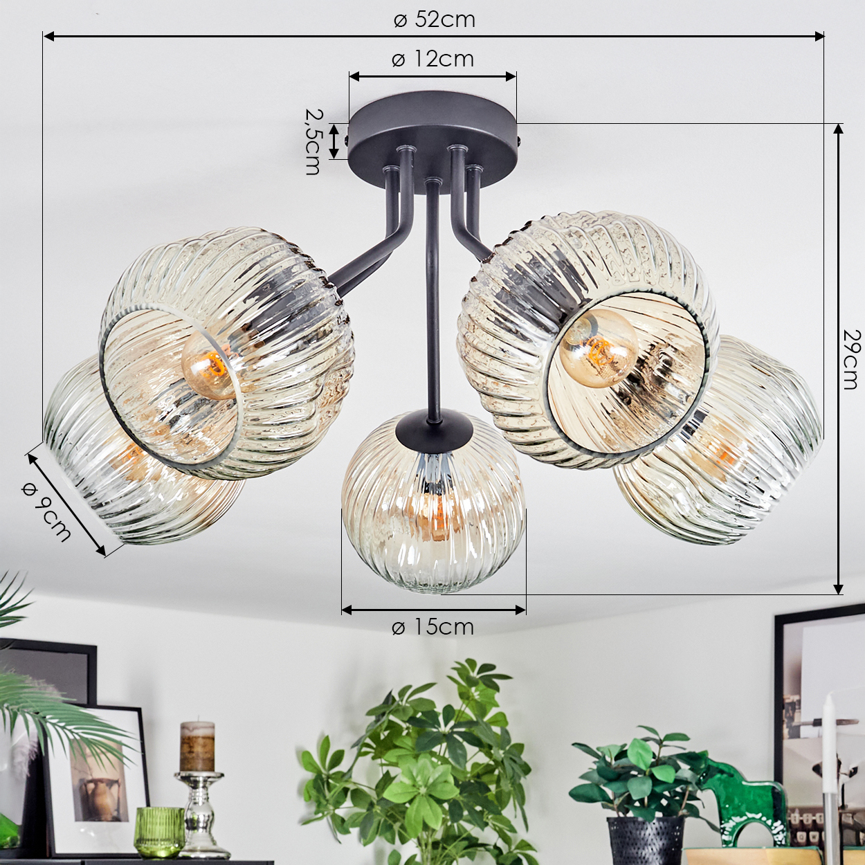 Eine moderne Deckenlampe mit fünf Glaskugeln, schwarzer Halterung und Maßen. Grüne Pflanzen und Bilderrahmen sind unten sichtbar.