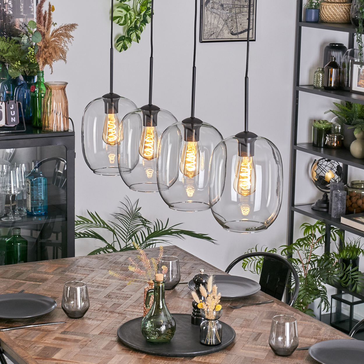 Esszimmer mit Holztisch, Glaslampen mit Glühbirnen, Geschirr, Gläsern, Pflanzen und einem schwarzen Metallregal mit Dekorationen.