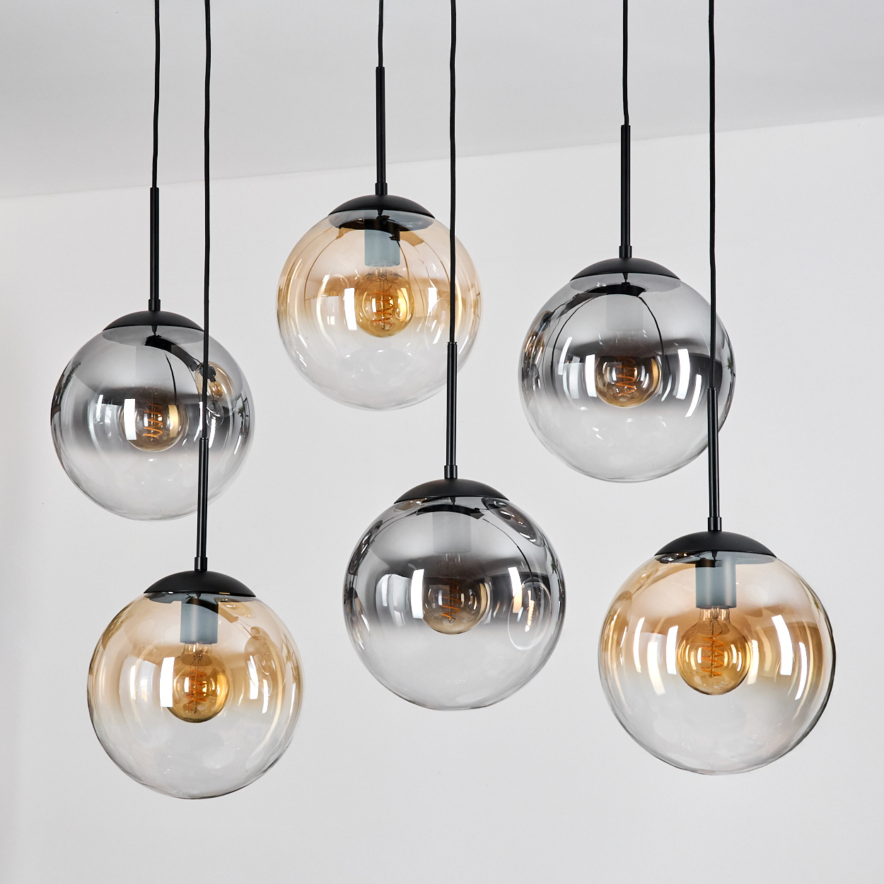 Sechs moderne Pendelleuchten mit Glaskugeln, einige bernsteinfarben, einige grau, mit sichtbaren Glühbirnen. Schwarze Kabel vor weißer Decke.