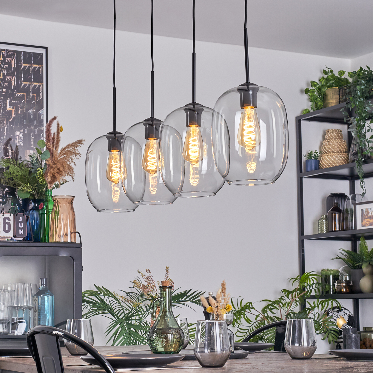 Esszimmer mit vier Glashängelampen. Tisch gedeckt mit Gläsern und Tellern. Regale mit Pflanzen, Vasen und Dekoration im Hintergrund.