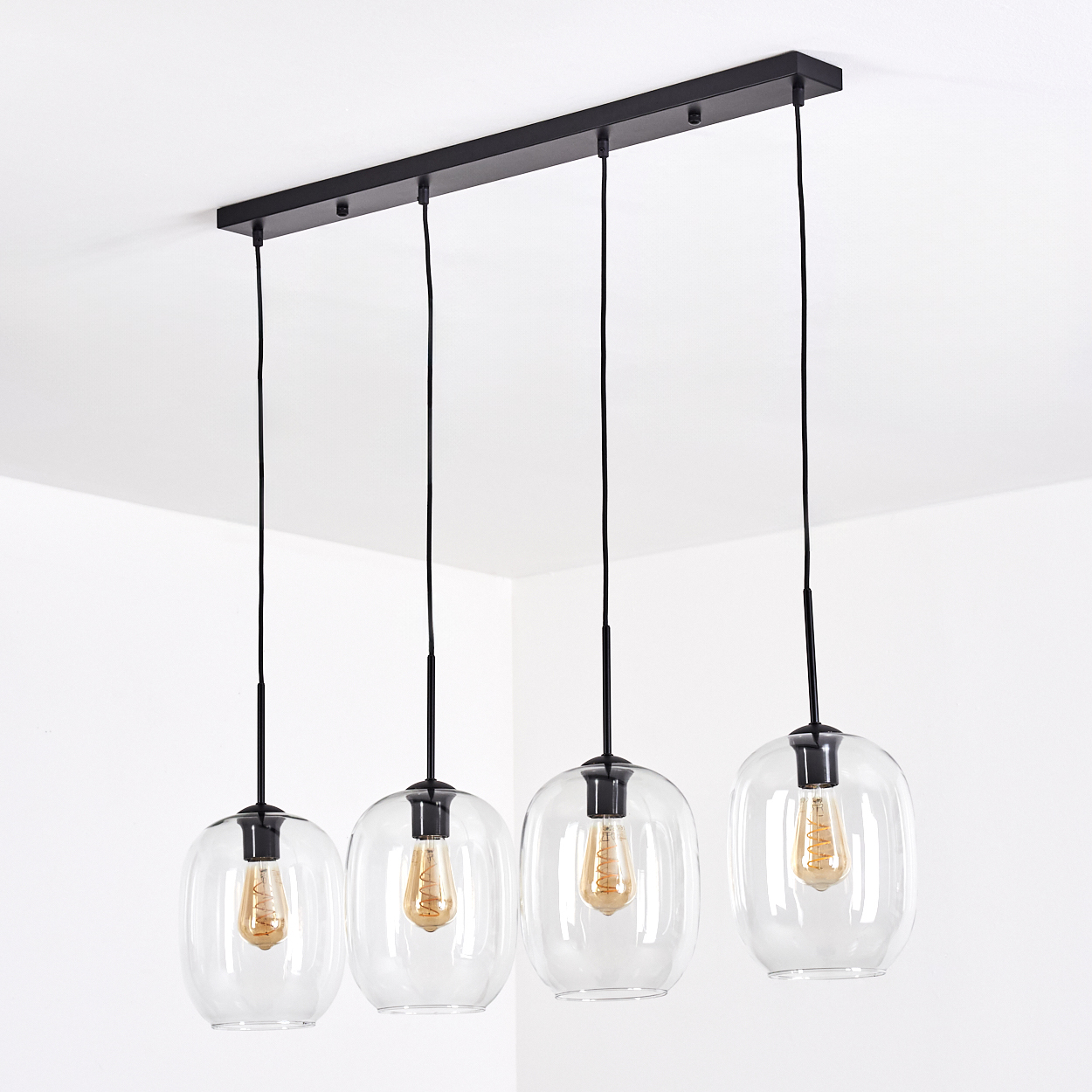 Vier Glas-Pendelleuchten mit schwarzer Hardware und sichtbaren Glühbirnen, hängen von einer schwarzen Deckenhalterung in einem weißen Raum.