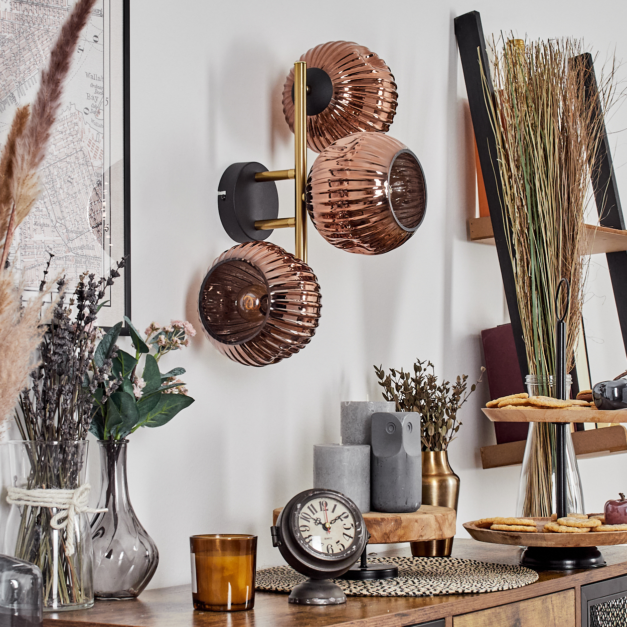Ein Raum mit einer kupferfarbenen Wandlampe, Vasen mit Blumen, einer Uhr, Kerzen und einem Holztablett mit Keksen neben einer schwarzen Leiter.