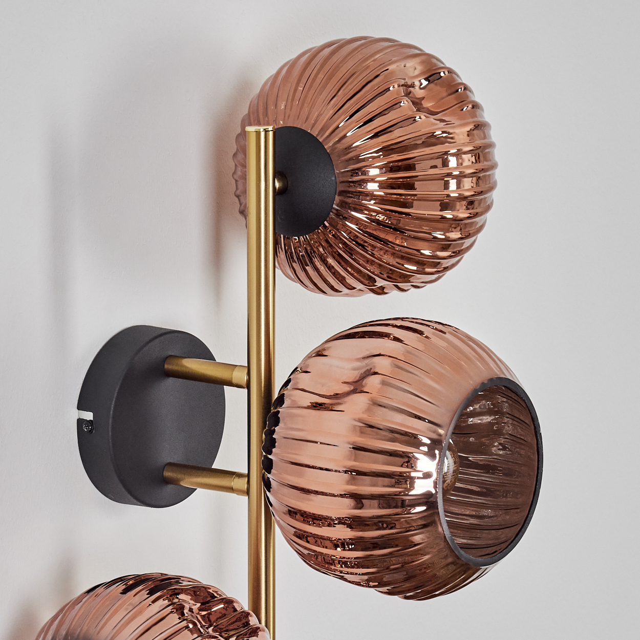 Eine moderne Wandlampe mit drei kupferfarbenen, gerippten Glasschirmen. Die Lampe hat goldfarbene Metallstützen und schwarze Montageplatten.