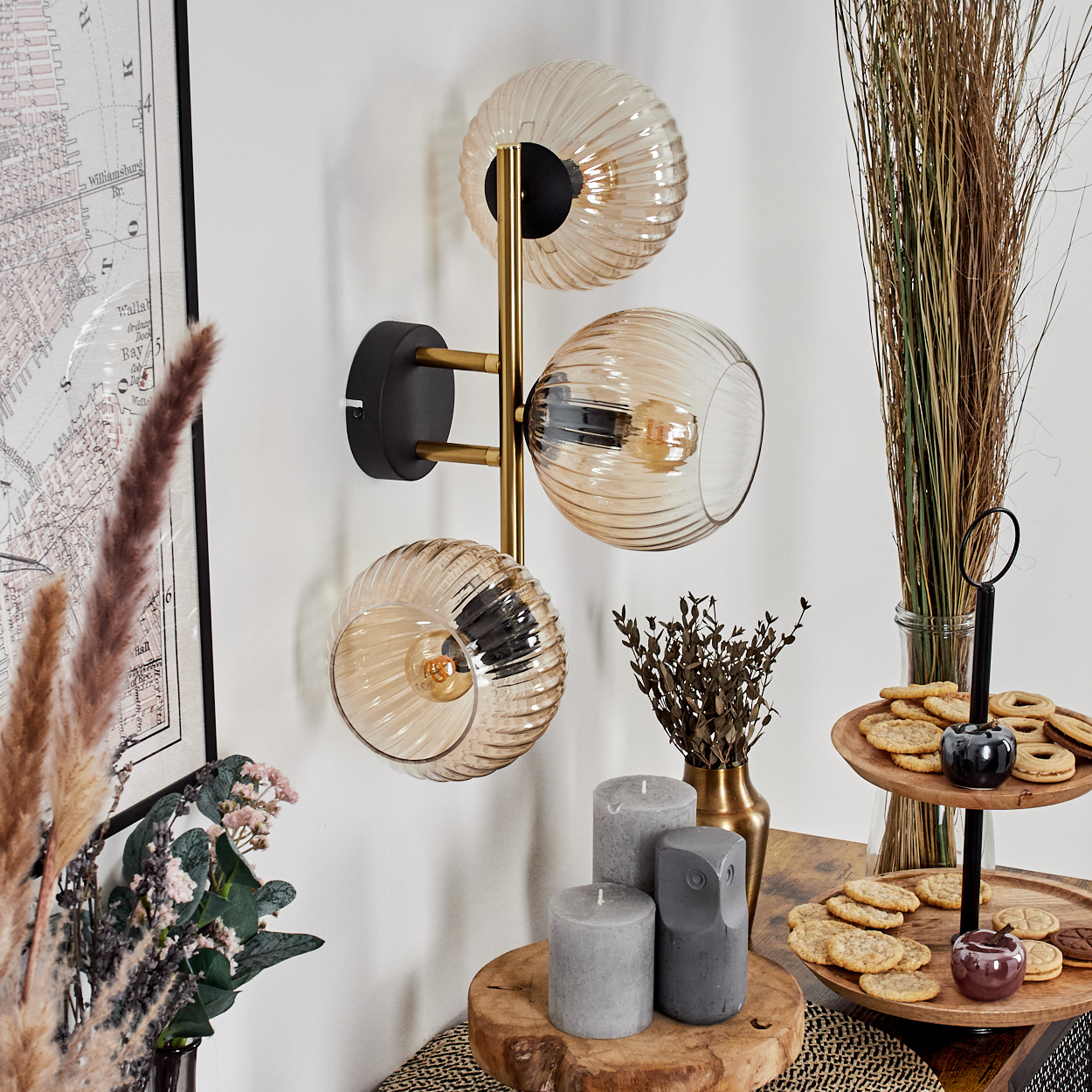 Eine Wandlampe mit drei Glaskugeln. Auf einem Holztisch stehen Kerzen, Kekse und getrocknete Pflanzen. Eine Karte ist sichtbar.