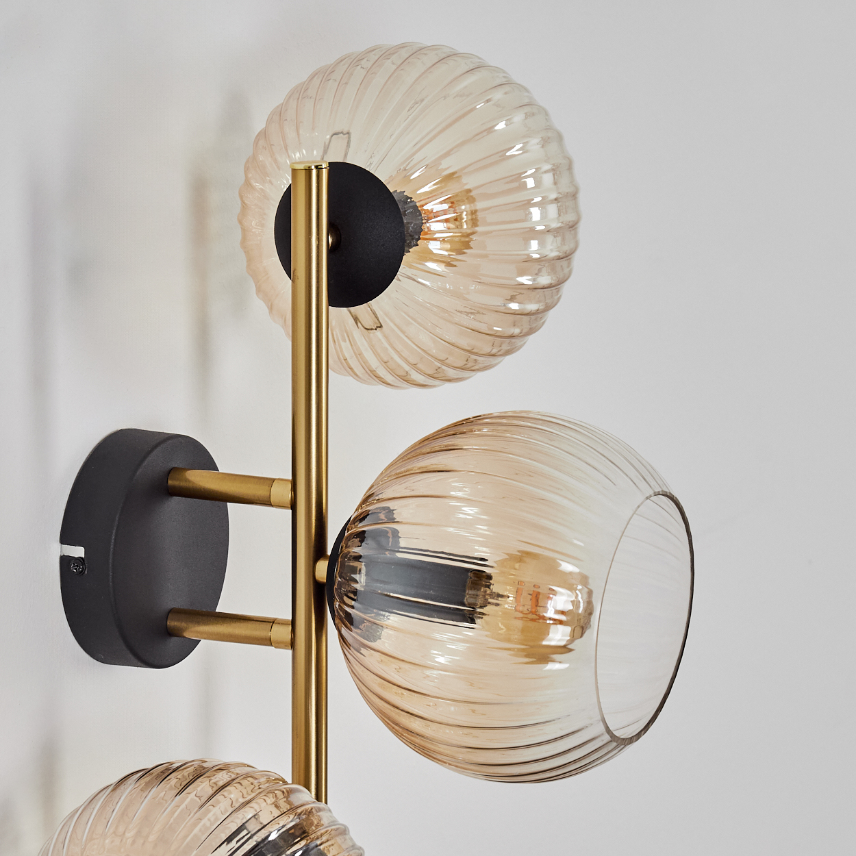 Eine moderne Wandlampe mit drei Glaskugeln. Die Kugeln sind bernsteinfarben und gerippt. Der Rahmen ist gold und schwarz. An einer weißen Wand montiert.