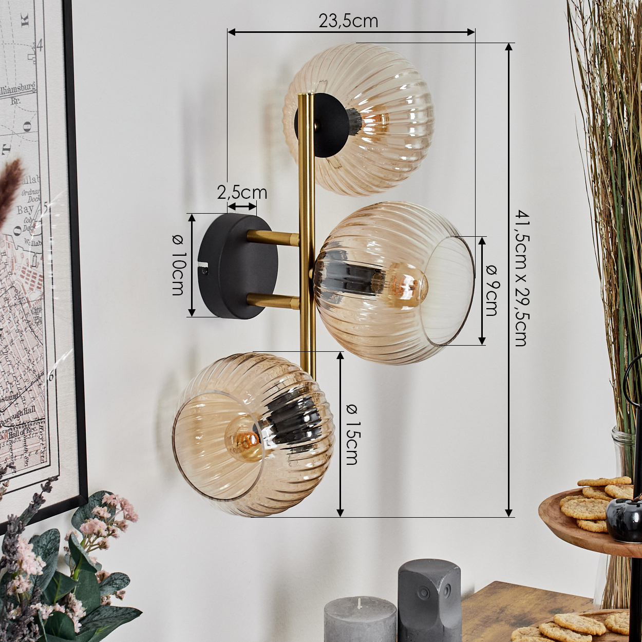 Eine Wandlampe mit drei Glaskugeln, goldenem Rahmen und schwarzem Sockel. Maße werden angezeigt. Andere Gegenstände sind im Hintergrund sichtbar.