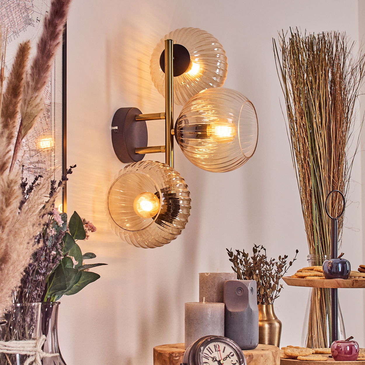Eine Wandleuchte mit drei Glaskugeln leuchtet. Getrocknete Pflanzen und Kerzen stehen auf einem Holztisch. Im Hintergrund ist eine Karte zu sehen.