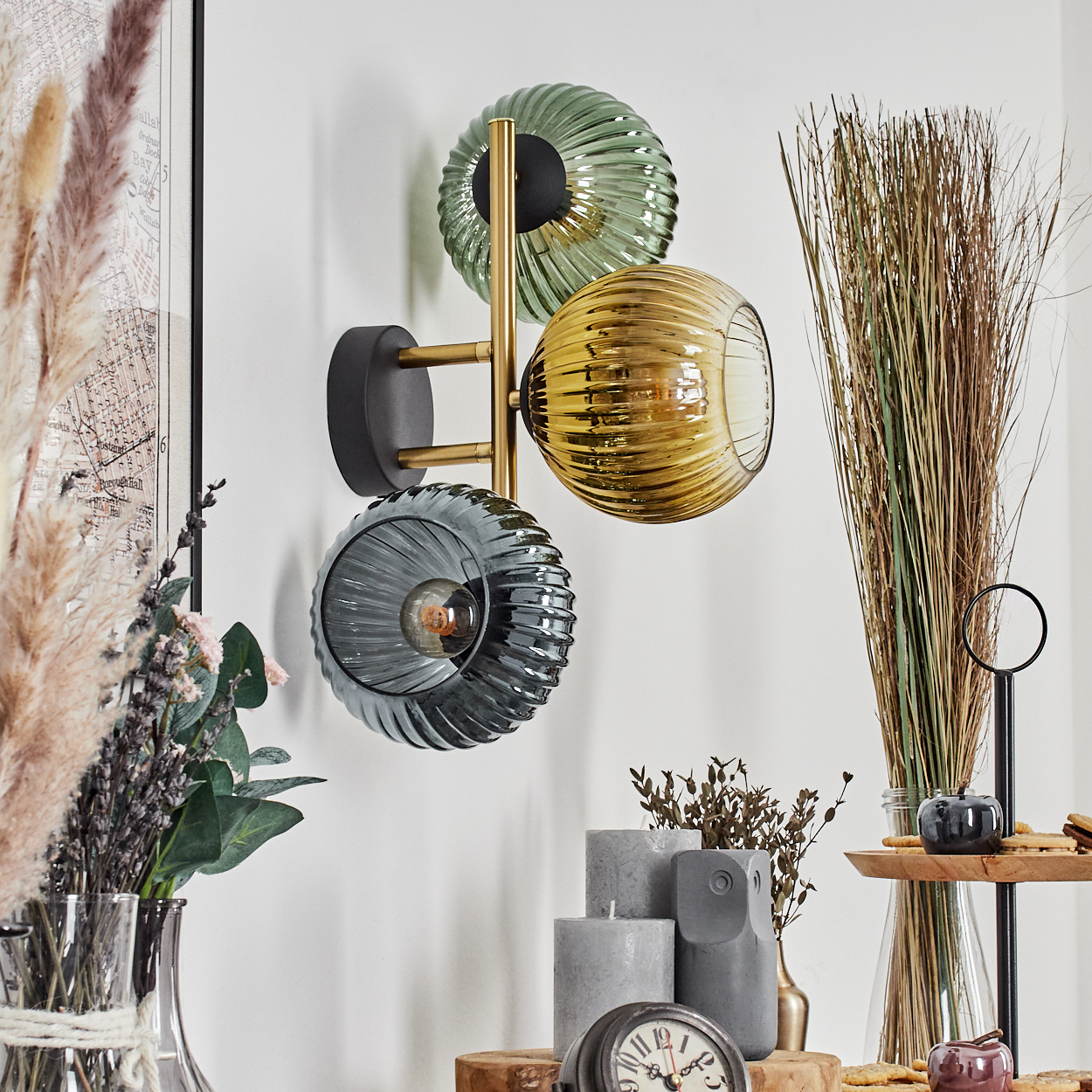 Eine Wandlampe mit drei Glaskugeln in Grün, Gelb und Grau. Dekorative Elemente wie Trockenpflanzen und Kerzen im Vordergrund.