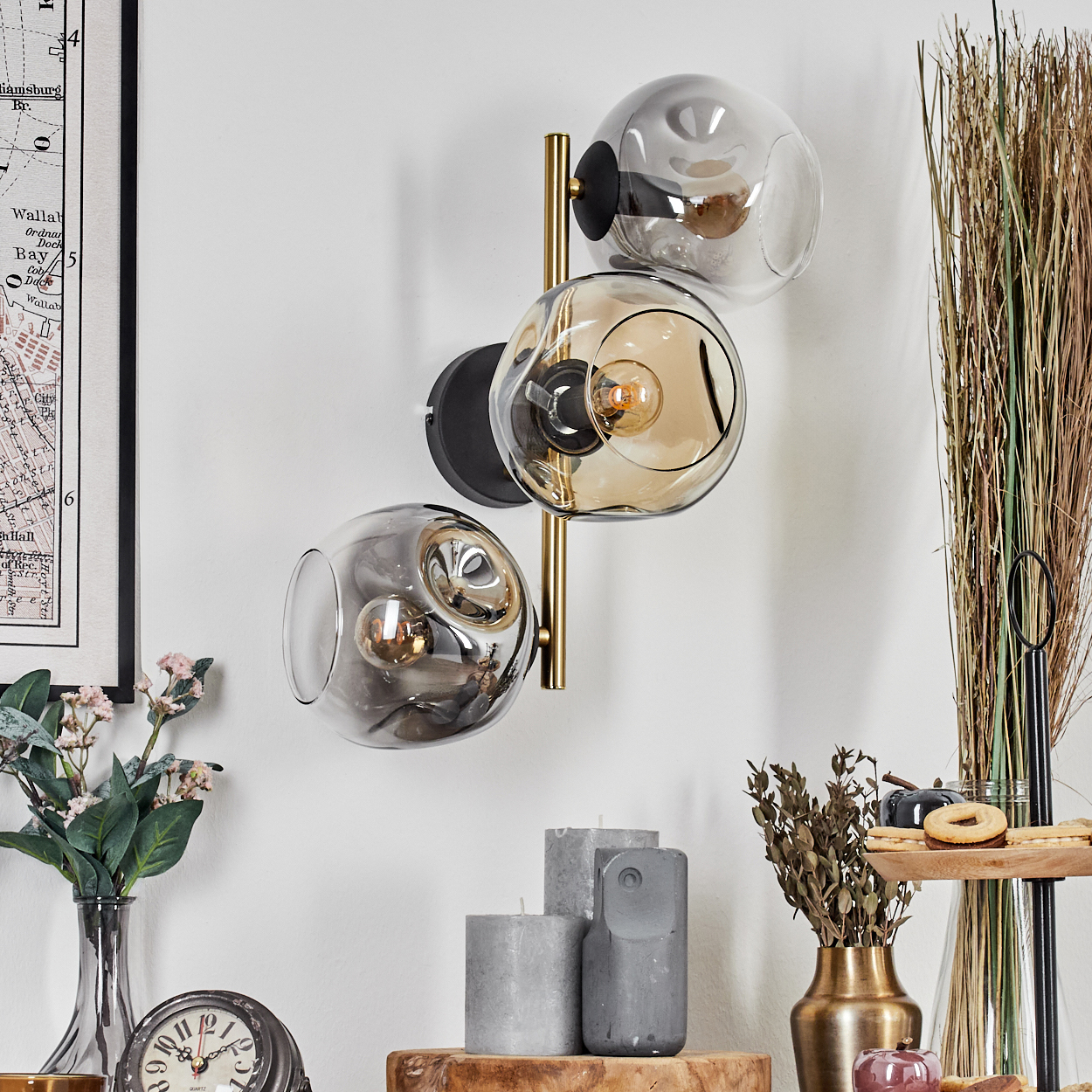 Eine moderne Wandlampe mit drei Glaskugeln. Verschiedene Dekorelemente, darunter Kerzen, Blumen und eine Uhr, sind sichtbar.