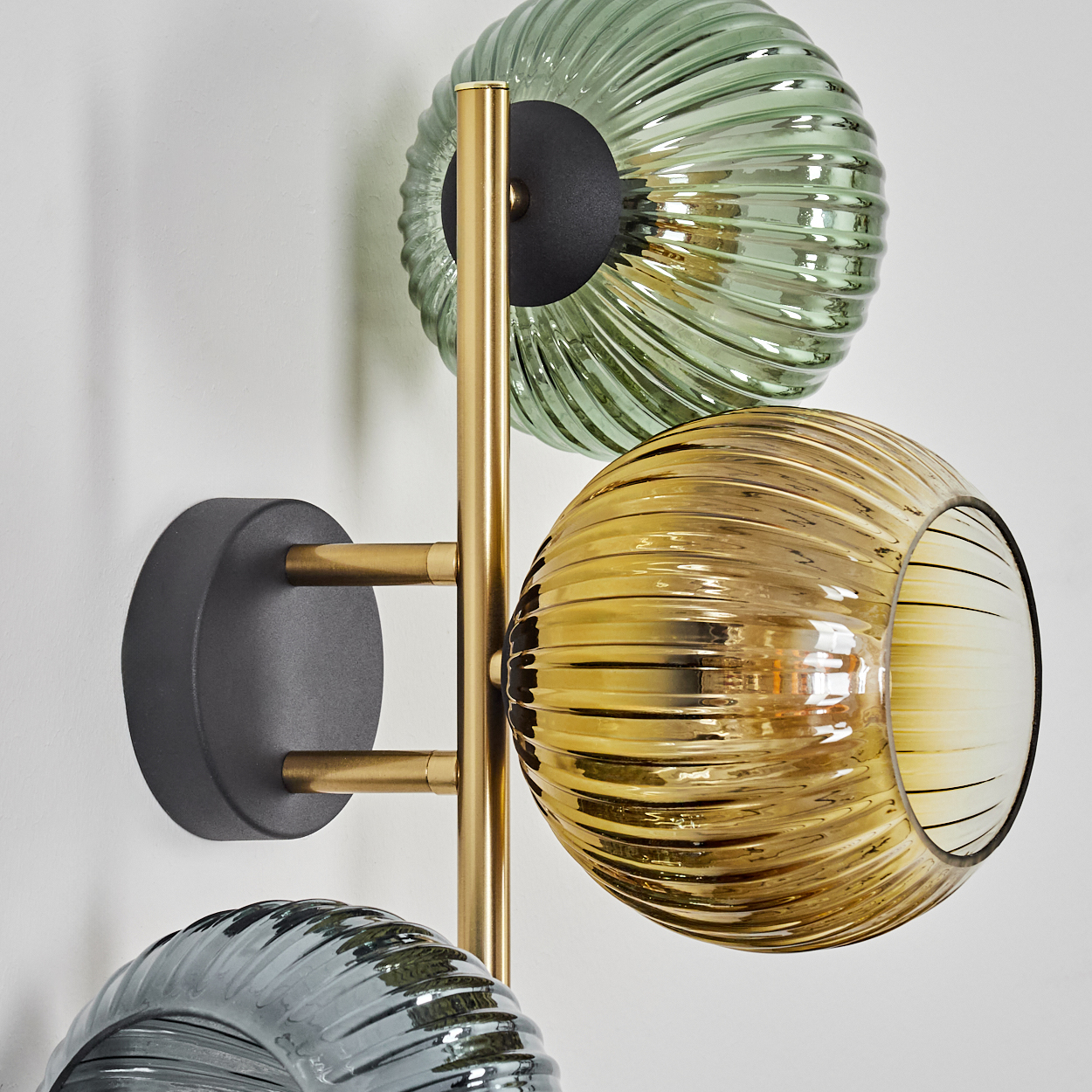 Eine moderne Wandlampe mit drei Glaskugeln in Grün, Gold und Grau. Die Lampe hat einen goldenen Rahmen und eine dunkelgraue Basis.