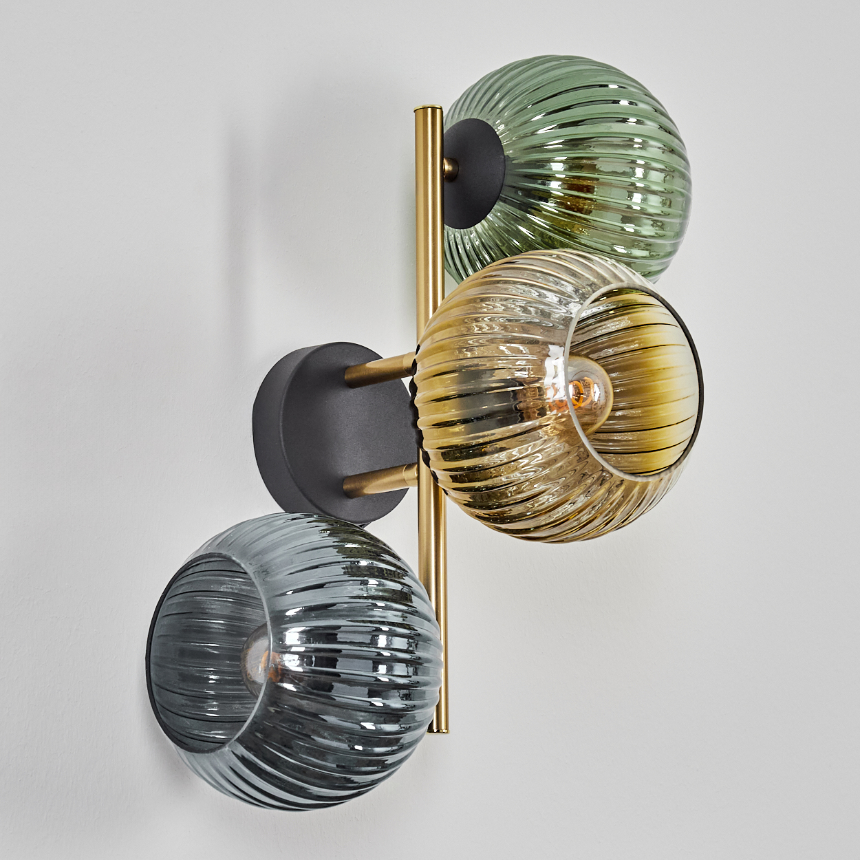 Eine Wandlampe mit drei Glaskugeln in Grün, Bernstein und Grau. Die Lampe hat eine goldene Stange und schwarze Montageplatten.