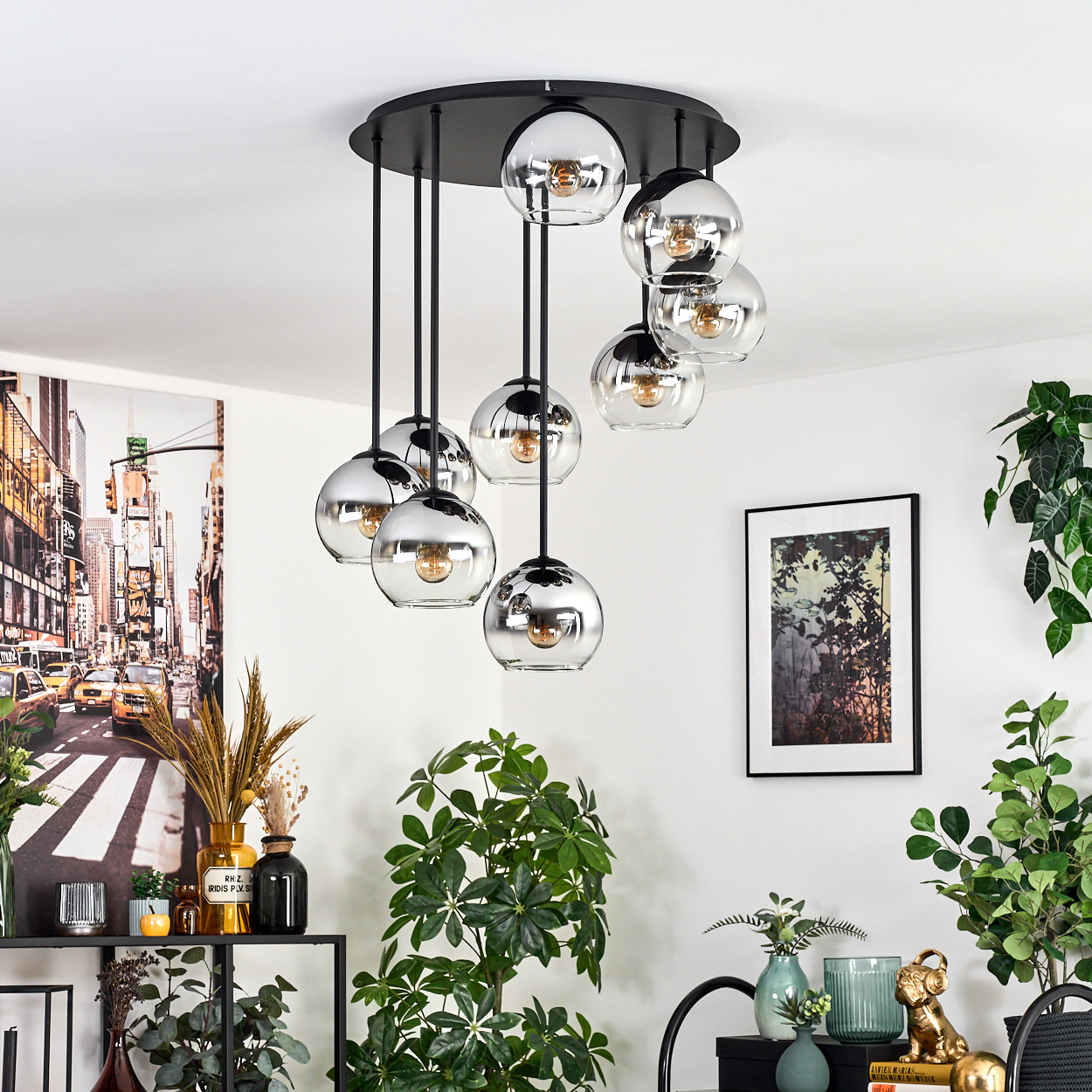 Eine moderne Deckenlampe mit kugelförmigen Chromlampen hängt von einer weißen Decke. Pflanzen und Kunstwerke schmücken die weiße Wand.