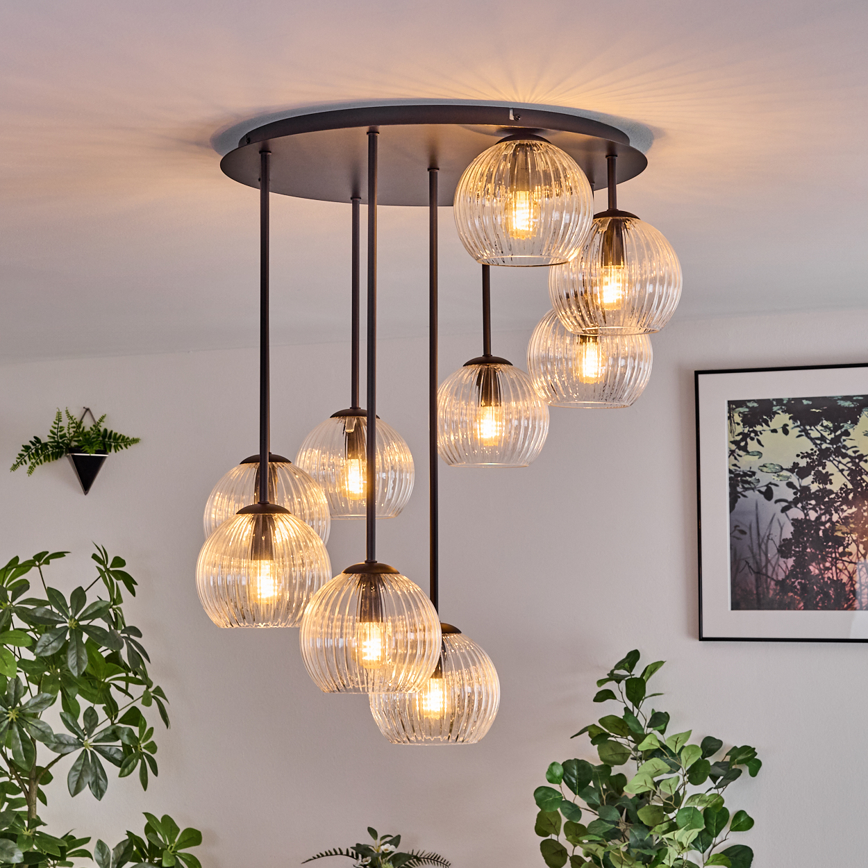Eine moderne Deckenlampe mit mehreren Glaskugellampen hängt von einer weißen Decke. Im Vordergrund sind grüne Pflanzen zu sehen.