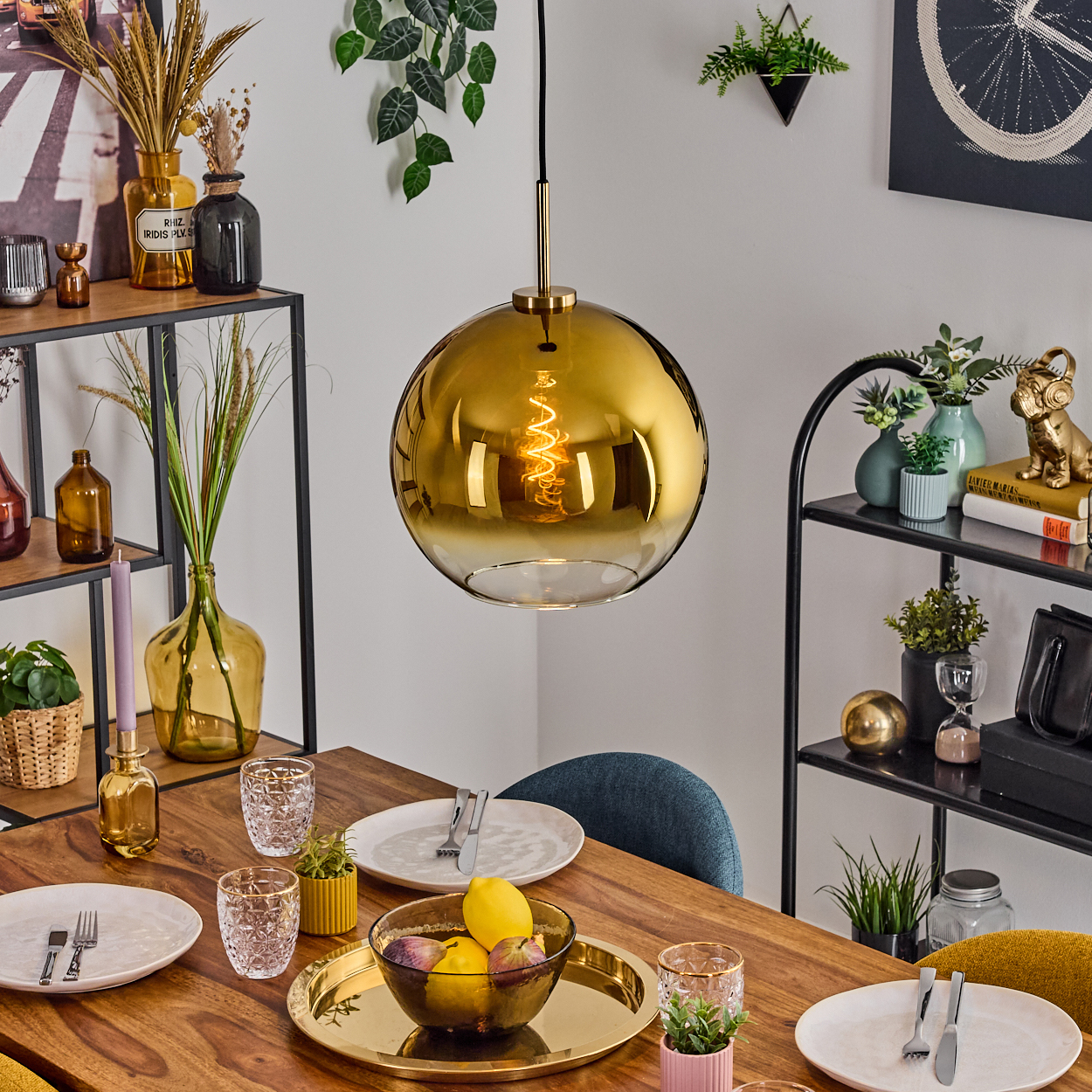 Esstisch mit Tellern, Gläsern und einer goldenen Pendelleuchte. Früchte in einer Schüssel. Regale mit Dekoration im Hintergrund.