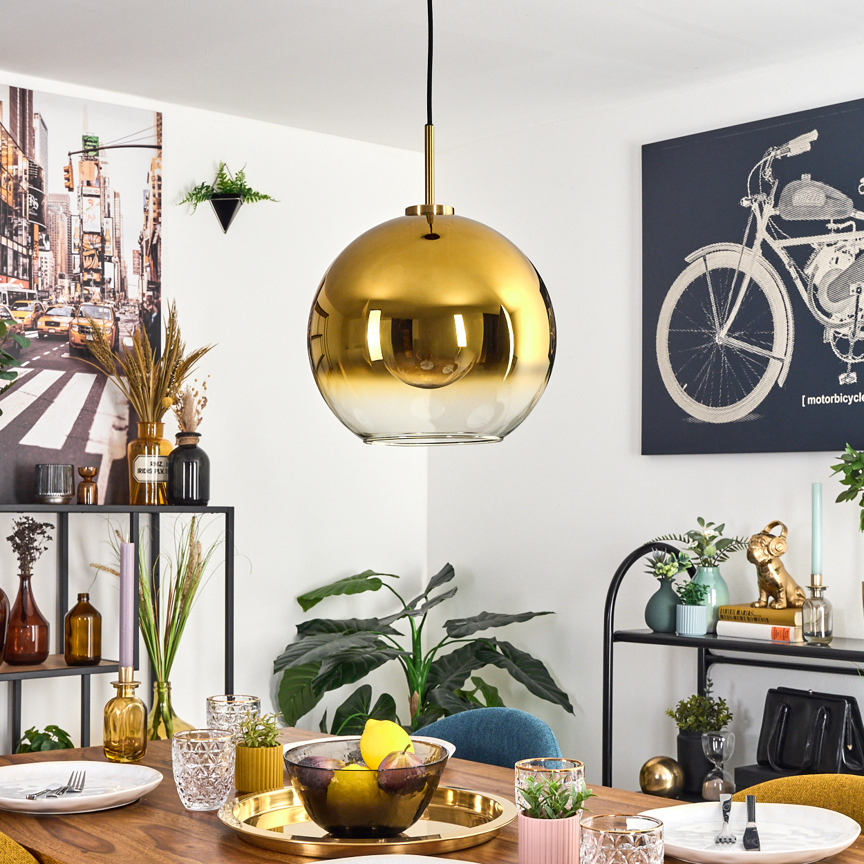 Ein Esszimmer mit einer goldenen Kugelleuchte. Tisch mit Tellern, Gläsern und einer Obstschale. Kunstwerke an den Wänden.