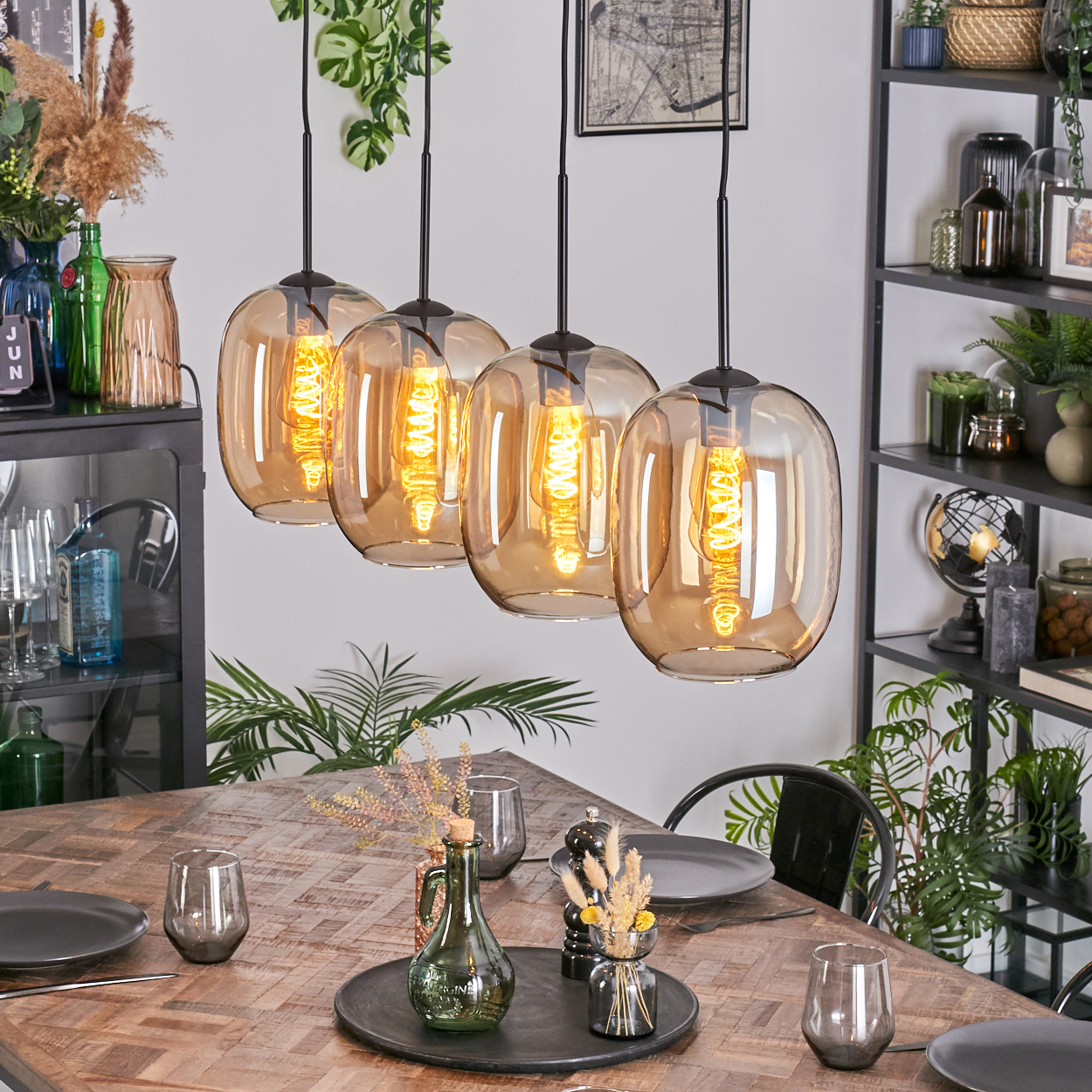 Esszimmer mit Holztisch, vier Pendelleuchten und verschiedenen Dekorationen. Pflanzen, Gläser und Teller sind sichtbar.