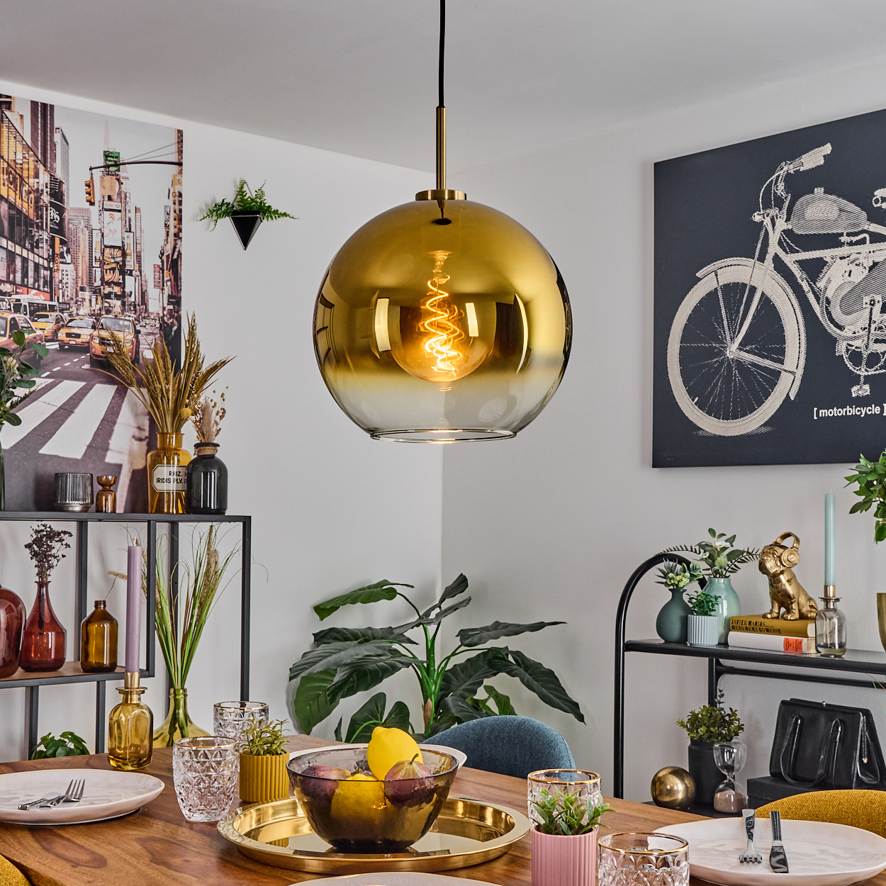 Esszimmer mit goldener Pendelleuchte, gedecktem Tisch und Obstschale. Kunst an den Wänden und Pflanzen.