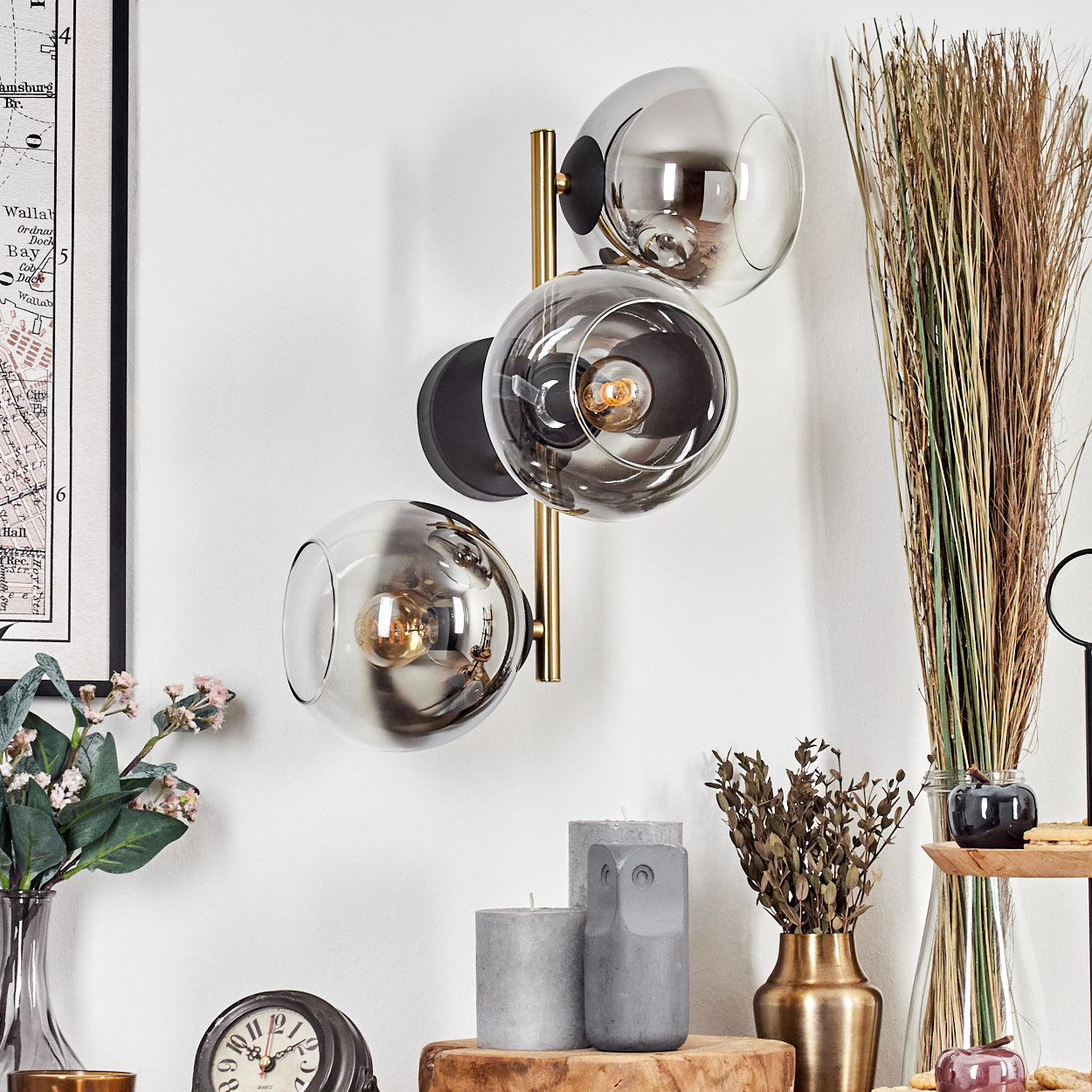 Eine Wandlampe mit drei Glaskugeln. Verschiedene Dekorationsartikel auf einer Holzoberfläche, darunter Kerzen, Vasen und Pflanzen.
