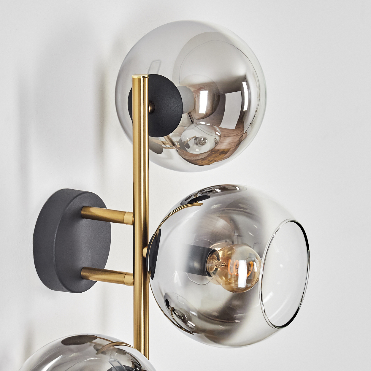 Eine moderne Wandlampe mit drei Glaskugel-Schirmen, Messingarmen und schwarzem Sockel vor weißer Wand.