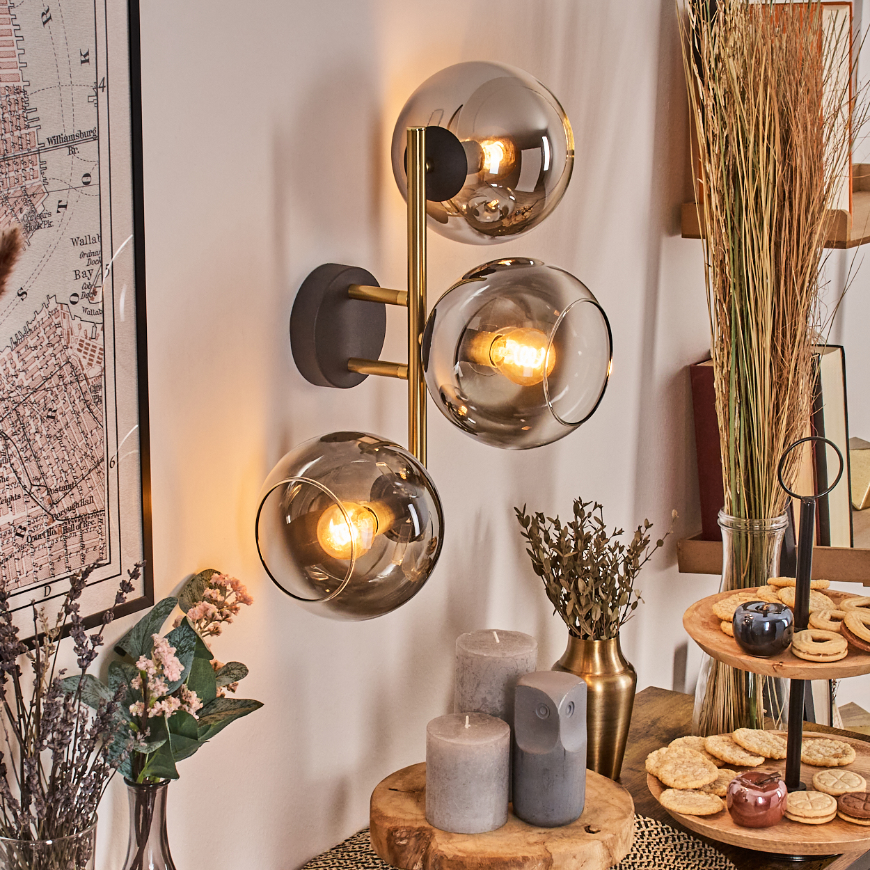 Eine Wandlampe mit drei Glaskugeln. Kerzen, Kekse und getrocknete Pflanzen stehen auf einem Tisch. Eine Karte hängt an der Wand.