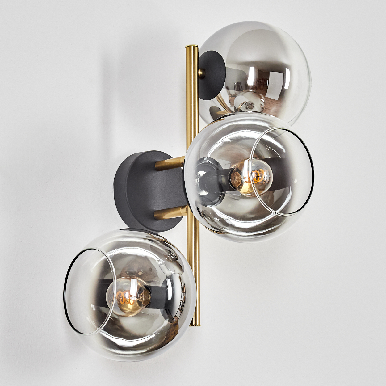 Eine moderne Wandlampe mit drei Glaskugel-Schirmen. Die Lampe hat ein goldfarbenes Metallgestell und ist an einer weißen Wand befestigt.