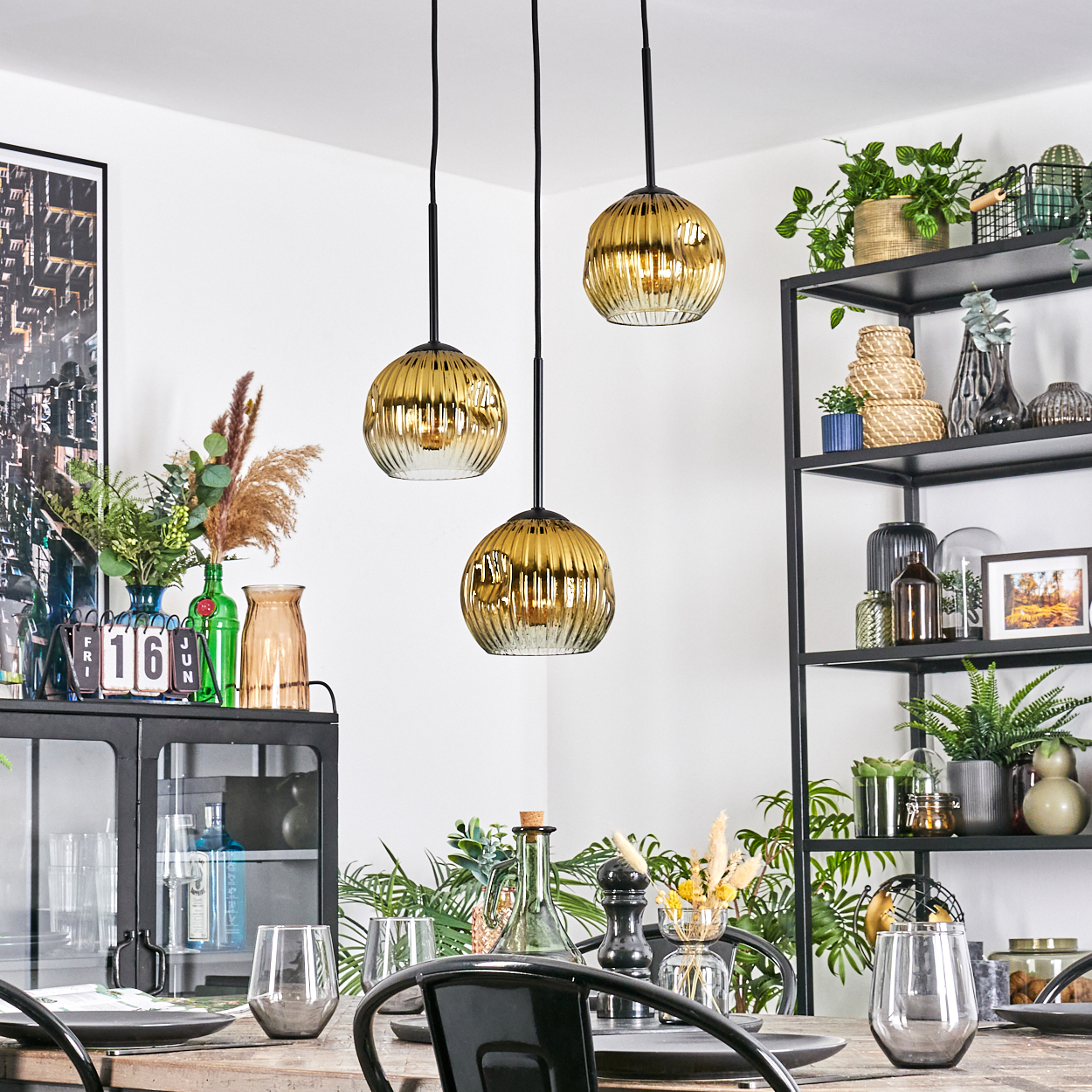 Esszimmer mit drei goldenen Pendelleuchten. Ein schwarzes Regal beherbergt Pflanzen und Dekoration. Tisch mit Gläsern und Tellern gedeckt.