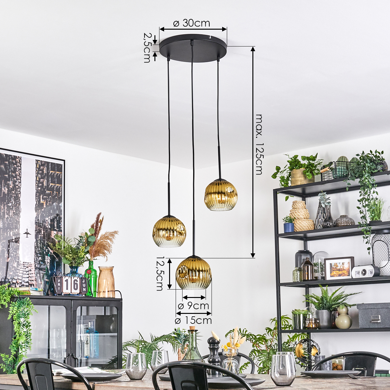 Drei goldene Pendelleuchten hängen über einem Esstisch. Rechts ein schwarzes Metallregal mit Pflanzen. Links ein Schrank und Kunst.