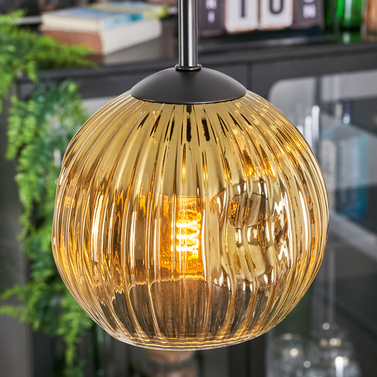 Eine goldene, gerippte Glas-Pendelleuchte ist beleuchtet und zeigt einen warmen Schein. Der Hintergrund umfasst Grün, Bücher und einen Schrank.