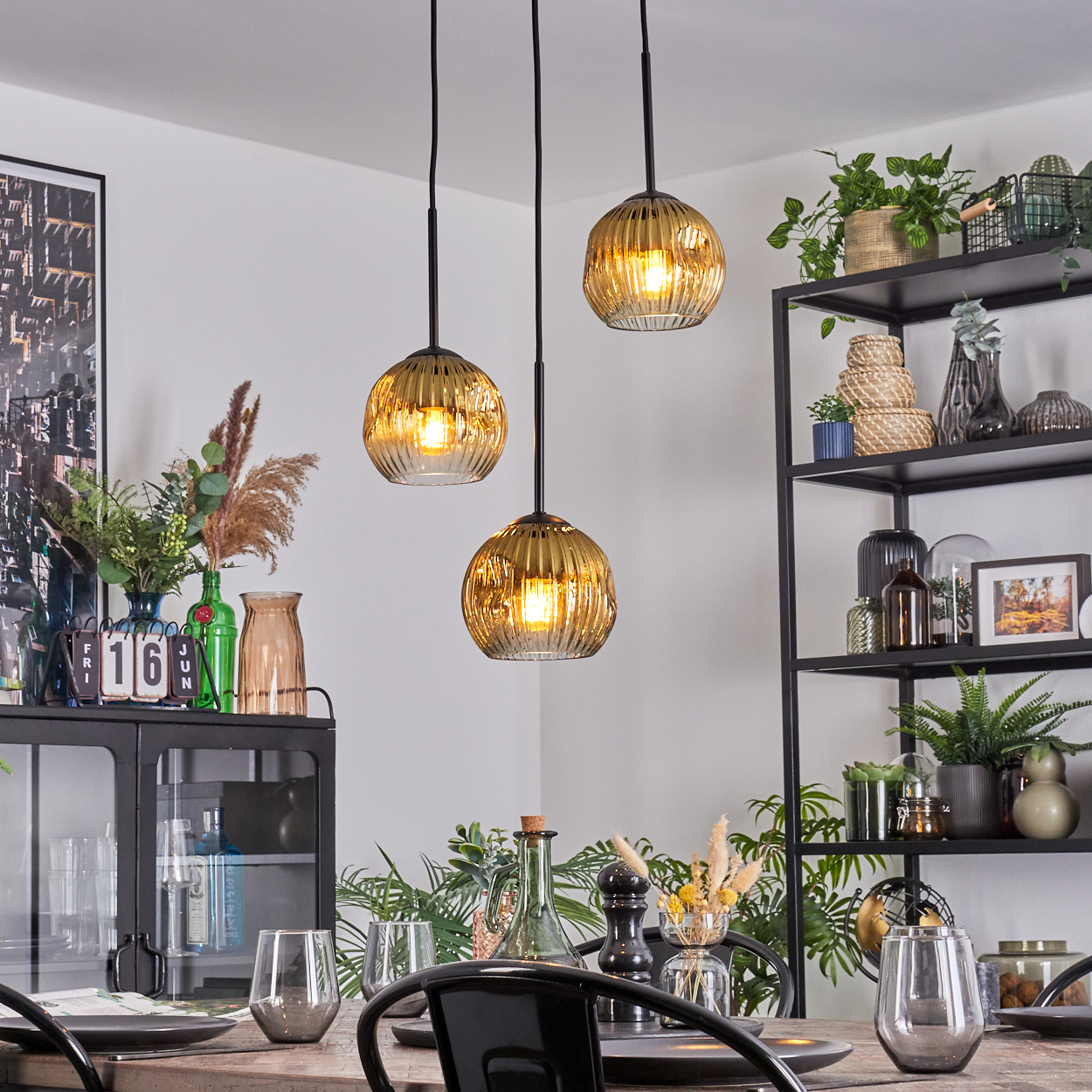 Esszimmer mit drei Hängelampen. Tisch mit Gläsern und Tellern. Regale mit Pflanzen und Dekoration.
