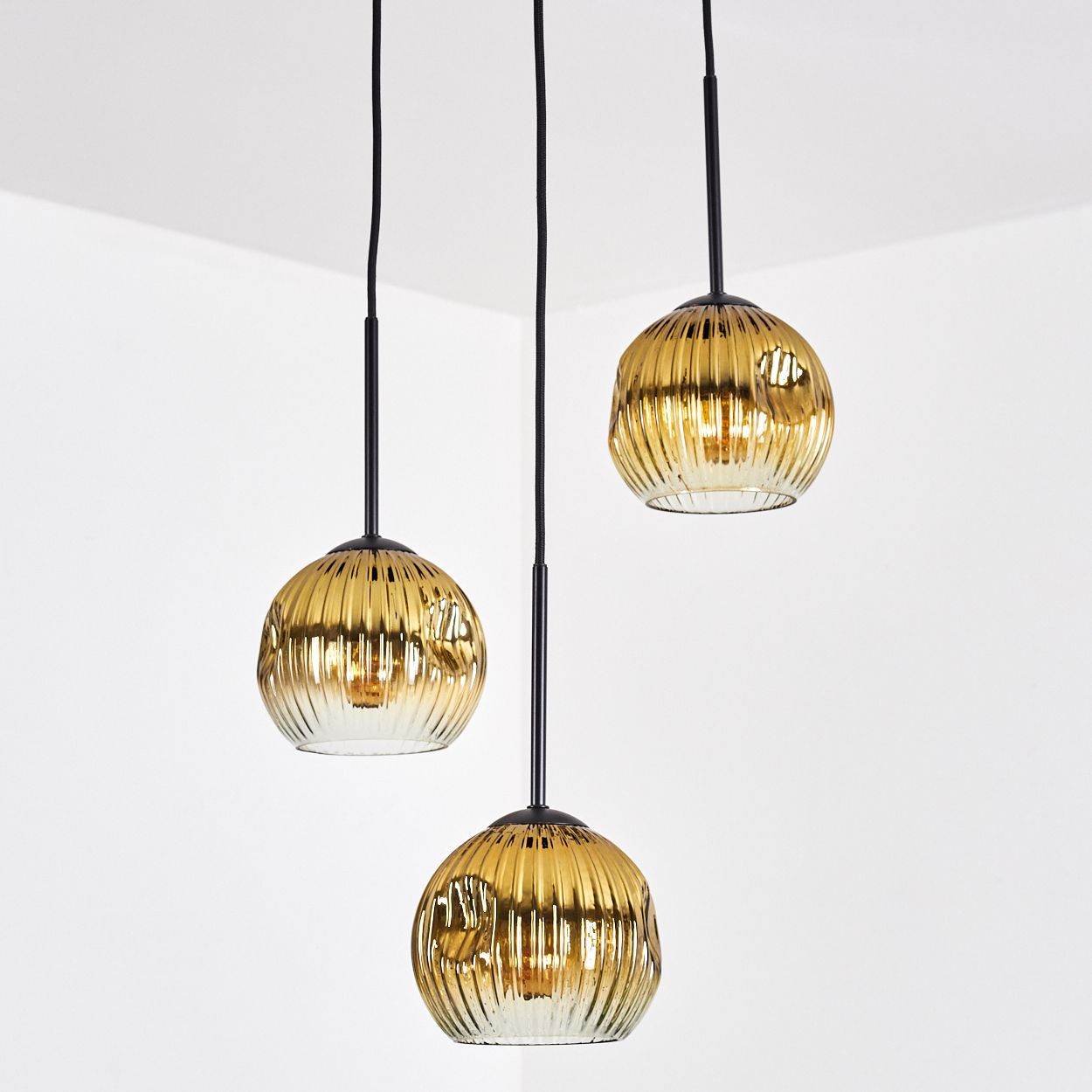 Drei goldene Pendelleuchten hängen an schwarzen Kabeln. Die Leuchten haben ein geripptes Glasdesign und befinden sich in einem weißen Raum.