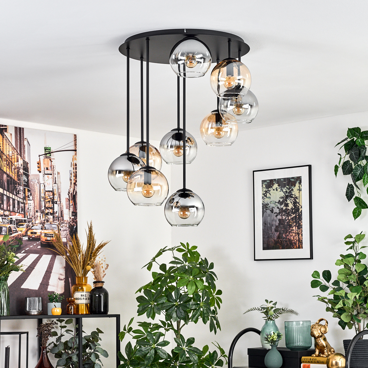 Eine moderne Hängelampe mit Glaskugeln hängt in einem Raum mit Pflanzen und Kunstwerken. Der Raum hat ein schwarzes Regal mit Vasen.