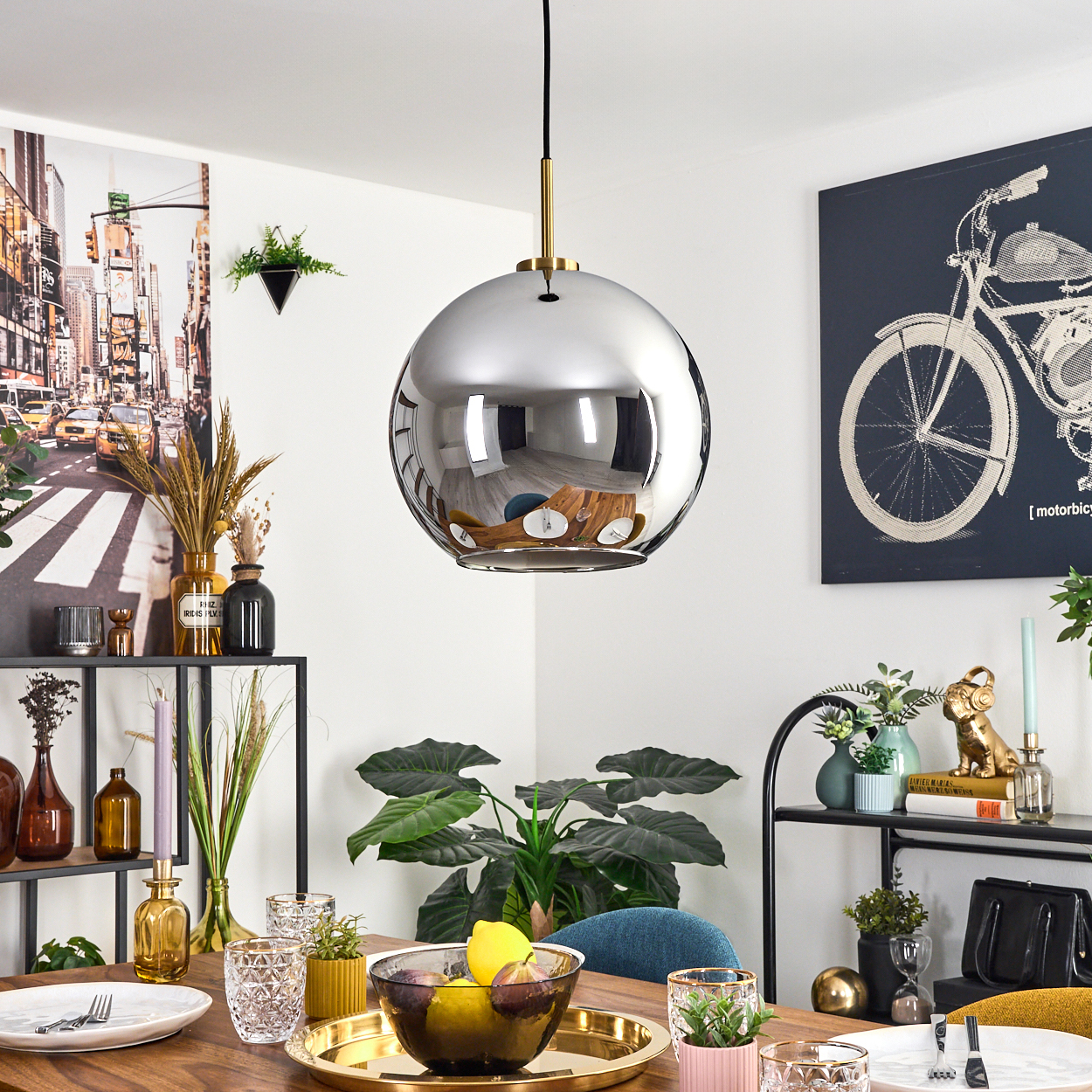 Ein Esszimmer mit einer reflektierenden Pendelleuchte. Ein Tisch ist mit Obst und Gläsern gedeckt. Kunstwerk eines Fahrrads an der Wand.