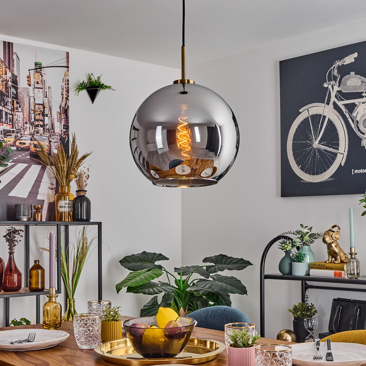 Esszimmer mit großer, reflektierender Pendelleuchte. Tisch mit Obst, Gläsern und goldener Platte. Kunstwerke an den Wänden.
