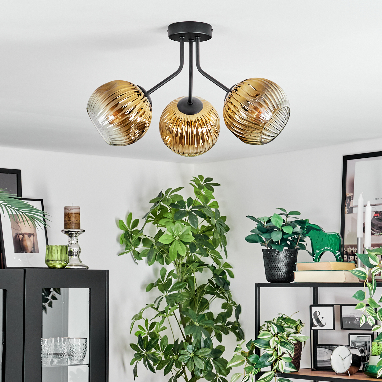 Eine moderne Deckenlampe mit drei goldenen Glasschirmen. Pflanzen und Dekorationen befinden sich im Hintergrund, darunter ein schwarzer Schrank und Regale.