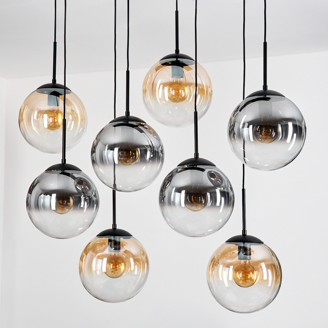 Acht Pendelleuchten mit Glaskugeln hängen von der Decke. Einige sind bernsteinfarben, andere klar mit Reflexen. Schwarze Armaturen und Drähte.