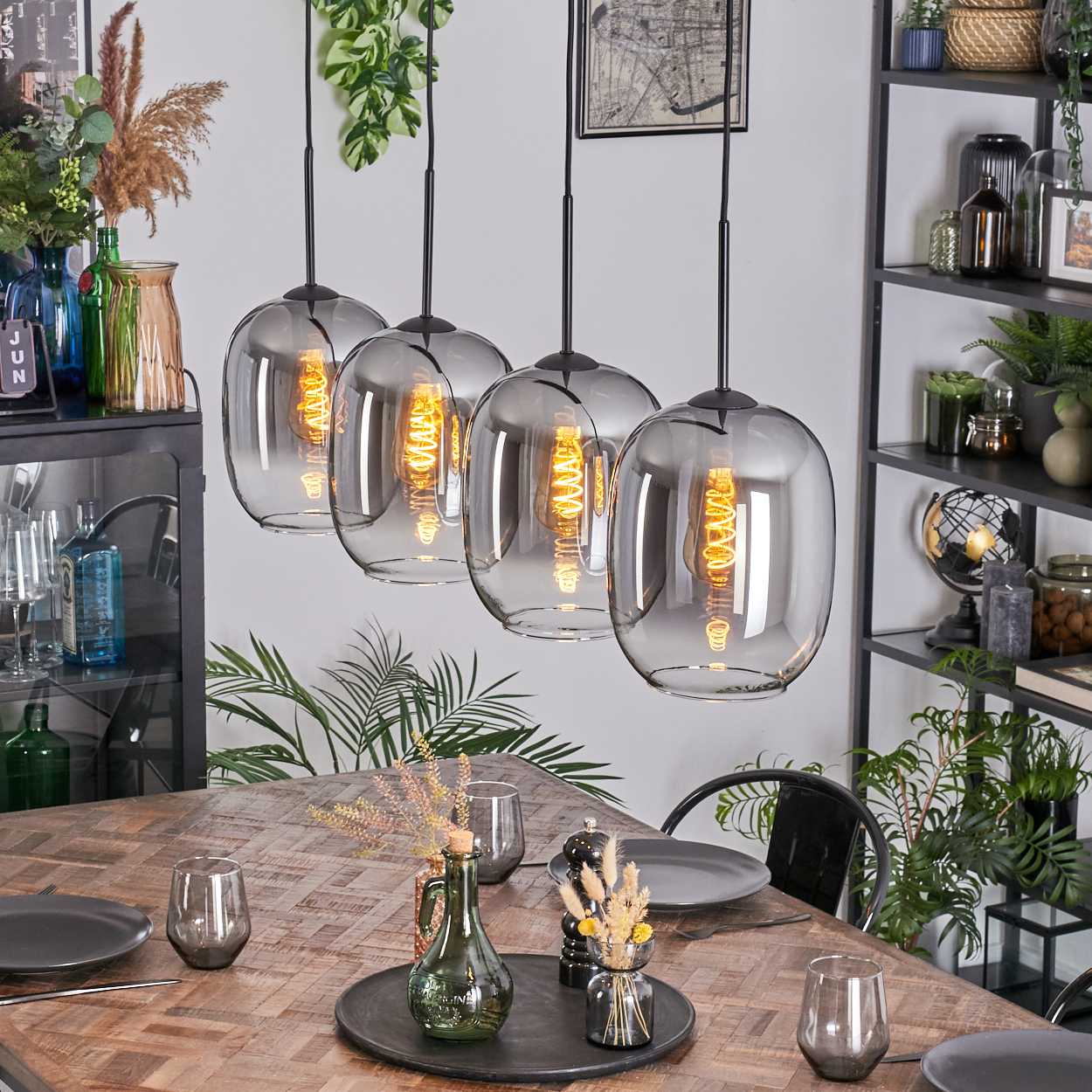 Esszimmer mit Holztisch, vier Pendelleuchten, Tellern, Gläsern und Dekoration. Regale mit Pflanzen und Dekoration.
