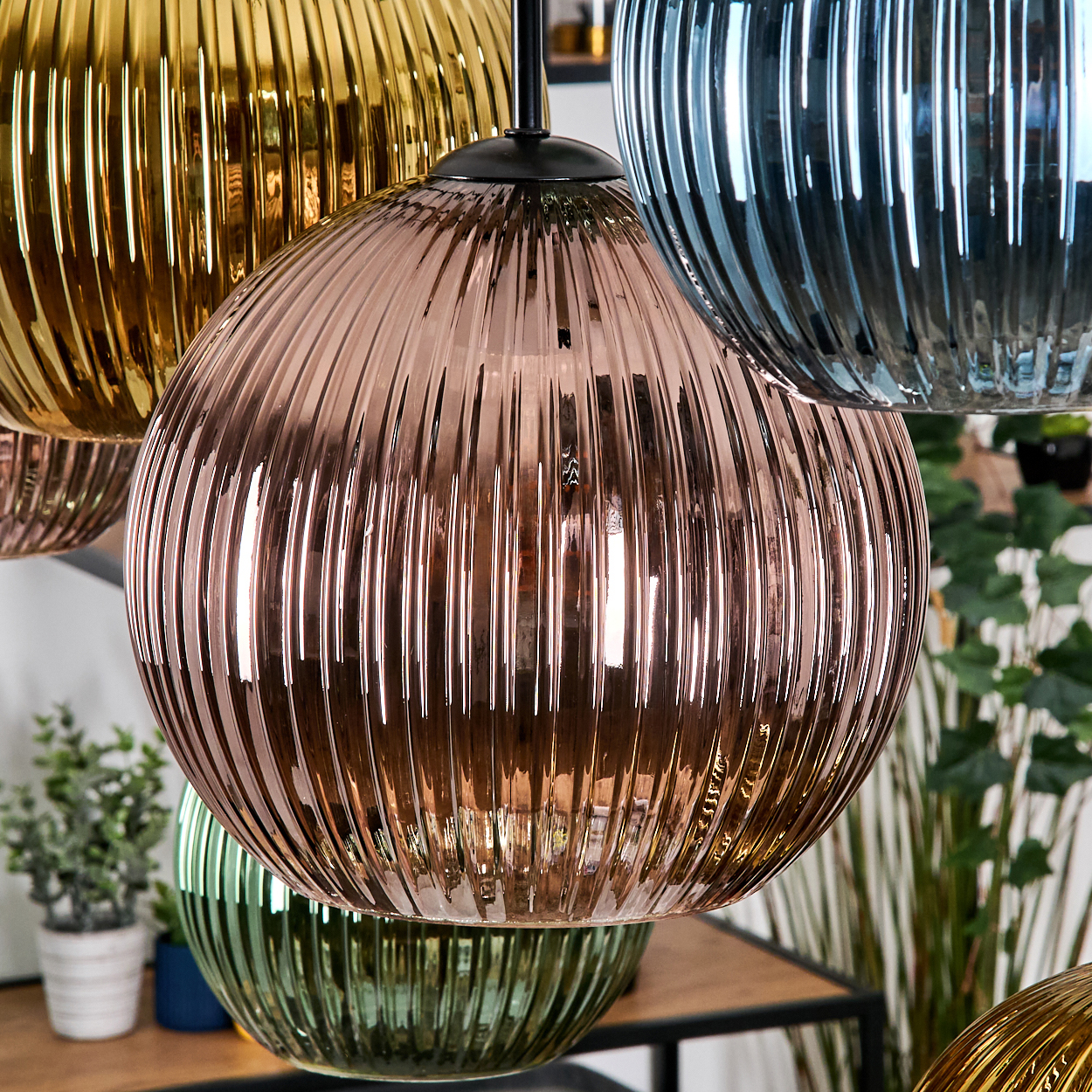 Nahaufnahme von hängenden Glaslampen in Gold, Rosa, Blau und Grün. Gerippte Textur. Pflanzen und Holzregal im unscharfen Hintergrund.