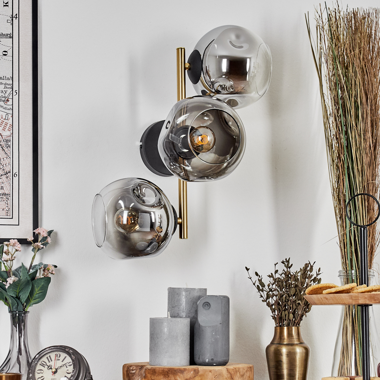 Eine Wandlampe mit drei Glaskugeln. Darunter Kerzen, eine Uhr und eine Vase mit Blumen. Trockene Pflanzen im Hintergrund.
