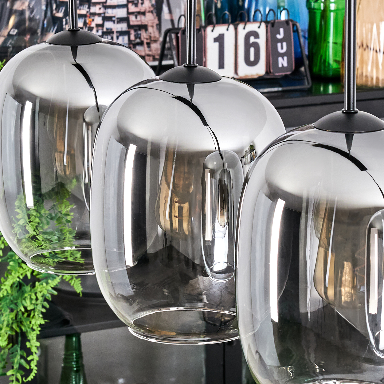Drei verspiegelte Glas-Pendelleuchten hängen. Ein Kalender zeigt den 16. Juni. Grüne Pflanzen und Flaschen im Hintergrund.