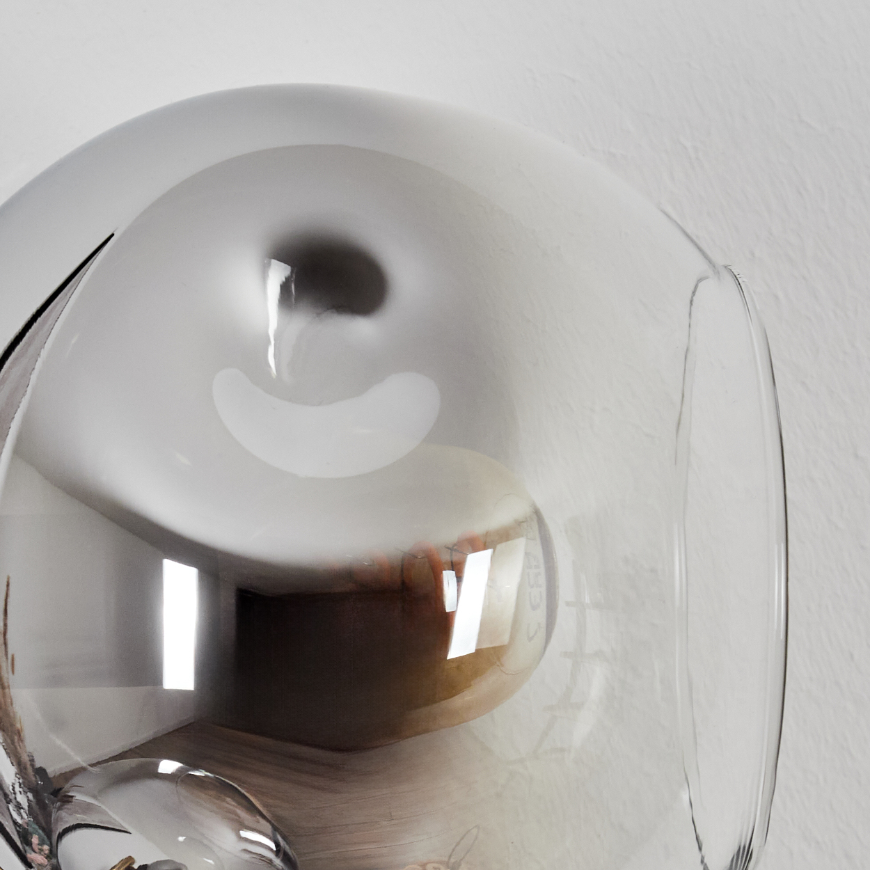 Nahaufnahme einer Glaslampe mit einer Glühbirne im Inneren. Die Lampe befindet sich an einer weißen Wand. Reflexionen sind auf dem Glas sichtbar.