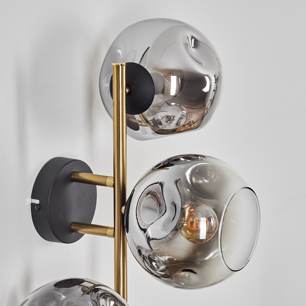 Eine moderne Wandlampe mit drei unterschiedlich großen Glaskugeln. Sie hat ein goldenes Gestell und einen schwarzen Sockel an einer weißen Wand.