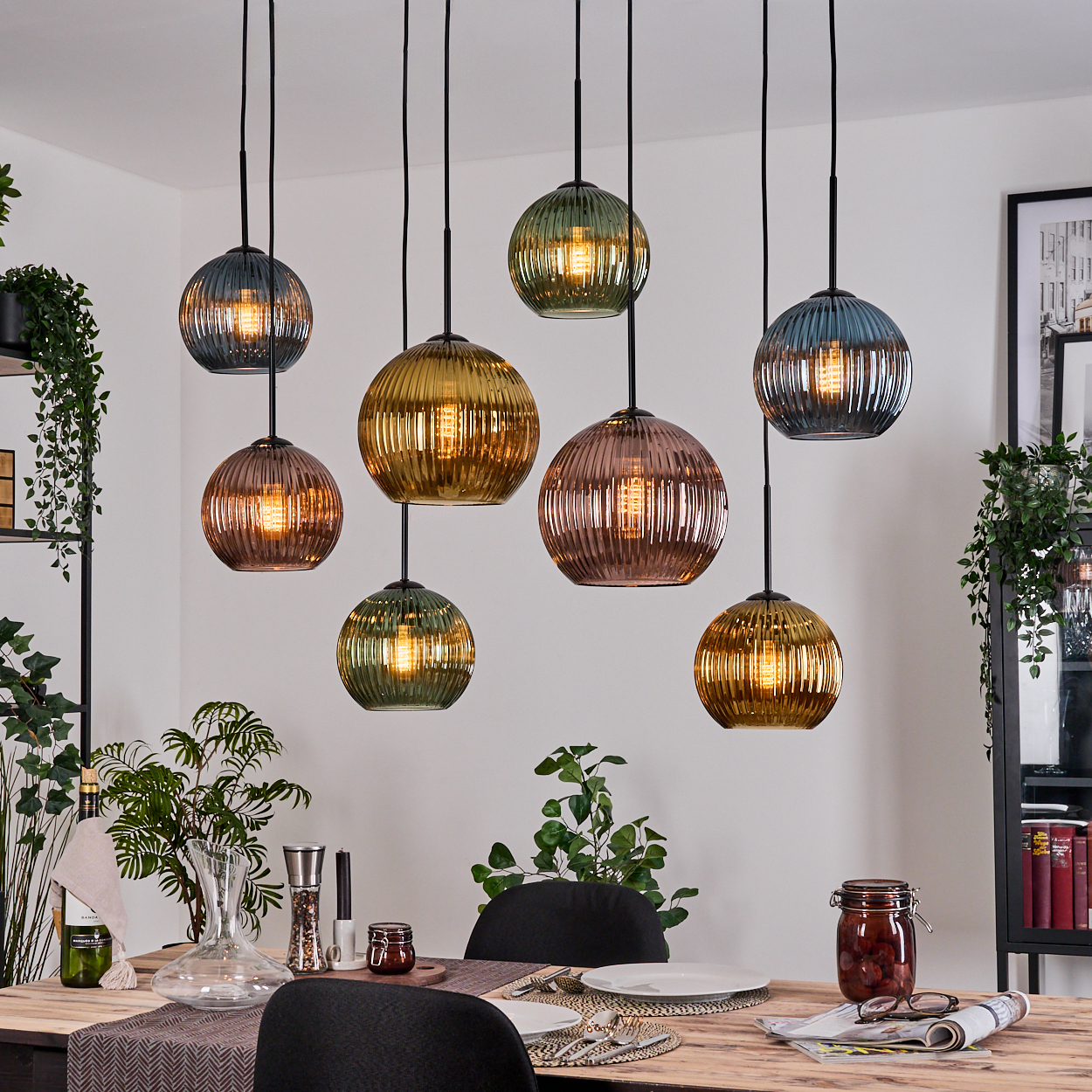 Esszimmer mit mehreren hängenden Glaskugelleuchten. Ein Tisch ist gedeckt mit Pflanzen, Geschirr und einem Glas mit Essen. Schwarze Stühle sind sichtbar.