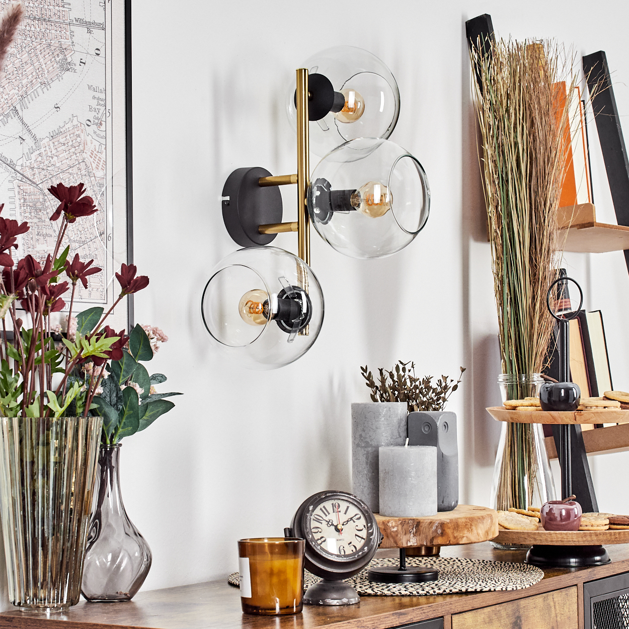 Eine Wandlampe mit drei Glaskugeln. Ein Tisch mit Blumen, Kerzen, einer Uhr und einem dekorativen Tablett. Im Hintergrund eine Karte und ein Regal.