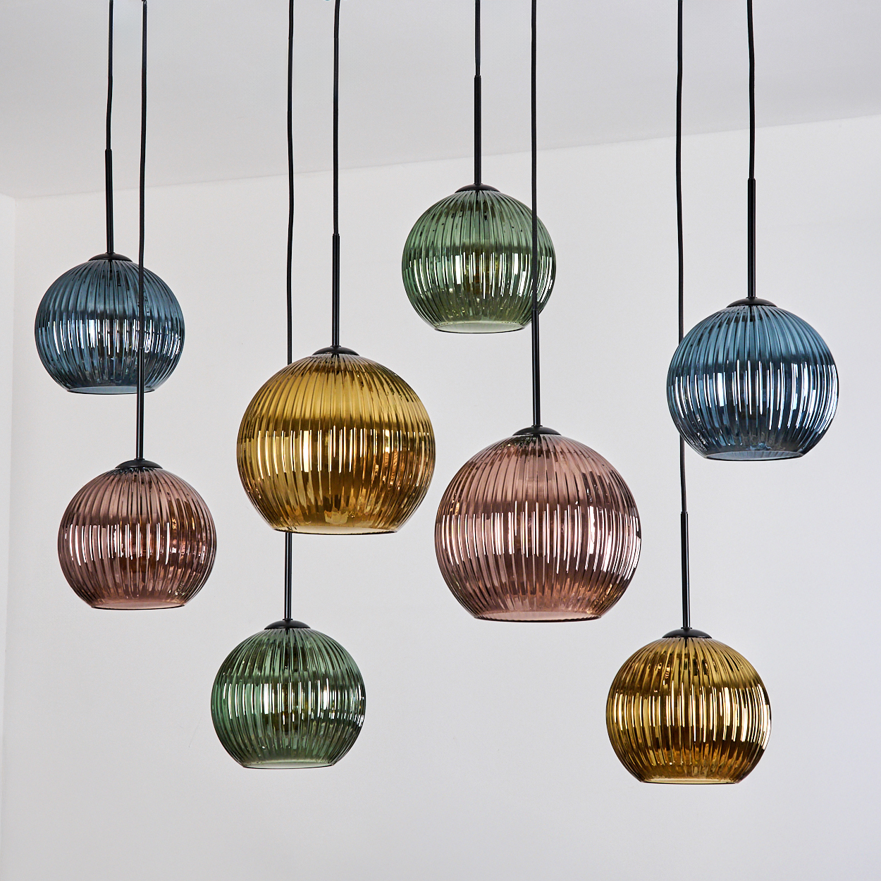 Mehrere Pendelleuchten hängen. Jede ist eine gerippte Glaskugel in verschiedenen Farben: blau, grün, gold und rosa, vor einem weißen Hintergrund.