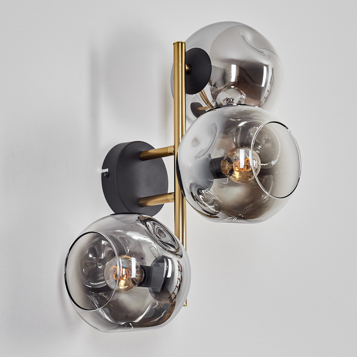 Eine moderne Wandlampe mit drei rauchigen Glaskugeln. Sie hat einen schwarzen Sockel, Messingarme und eine goldene Stange an einer weißen Wand.