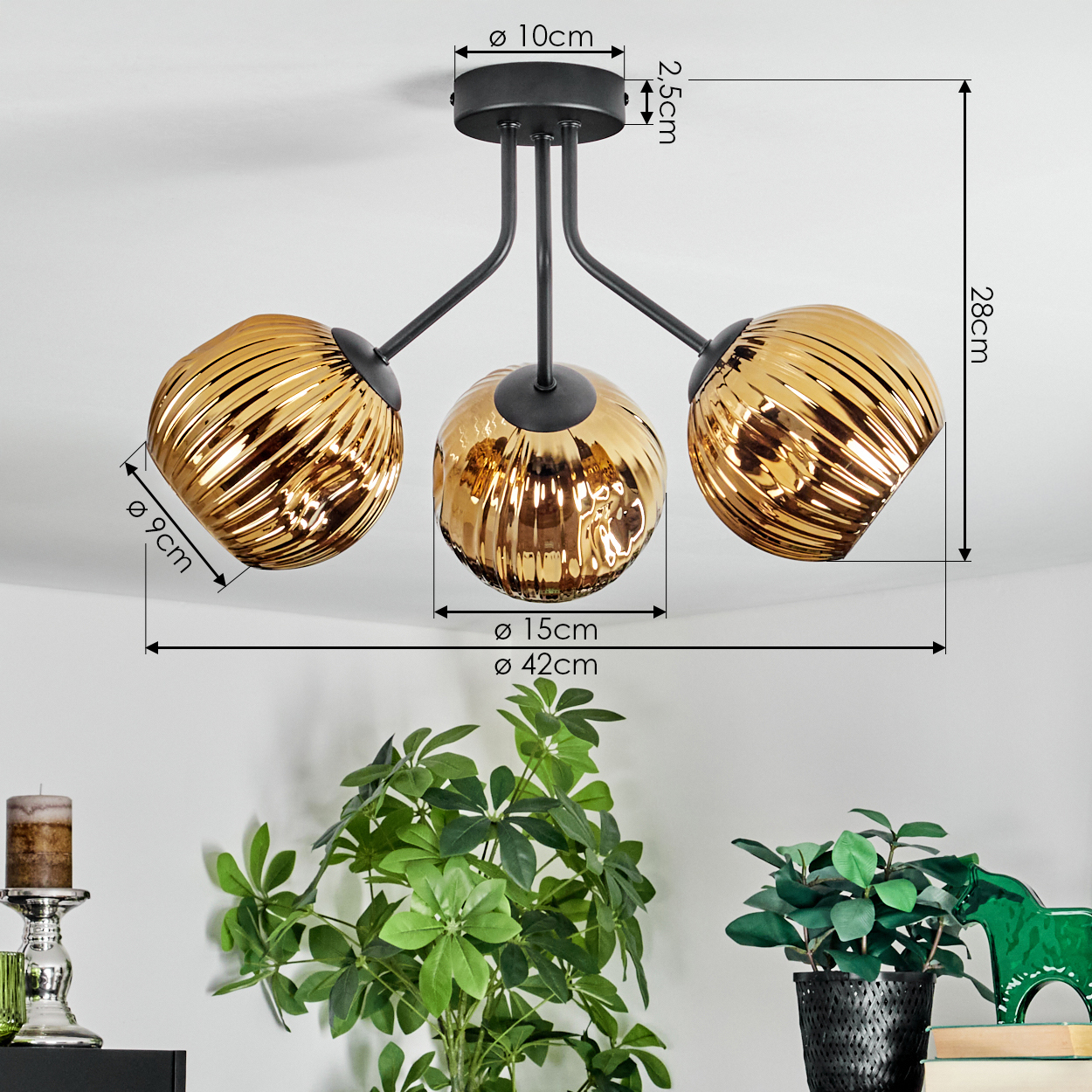 Moderne Deckenlampe mit drei goldenen Glasschirmen. Über grünen Pflanzen und einer Kerze auf einem Regal. Abmessungen sind angegeben.