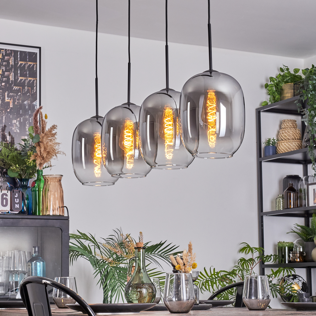 Vier Glas-Pendelleuchten über einem gedeckten Esstisch. Der Tisch ist mit Gläsern und Tellern gedeckt. Der Hintergrund umfasst Deko und Pflanzen.