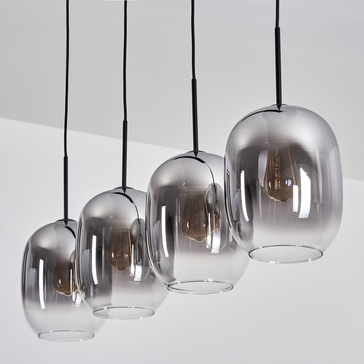 Vier Glas-Pendelleuchten, jede mit einer Glühbirne, hängen an schwarzen Kabeln von einer weißen Decke. Das Glas ist rauchgrau.