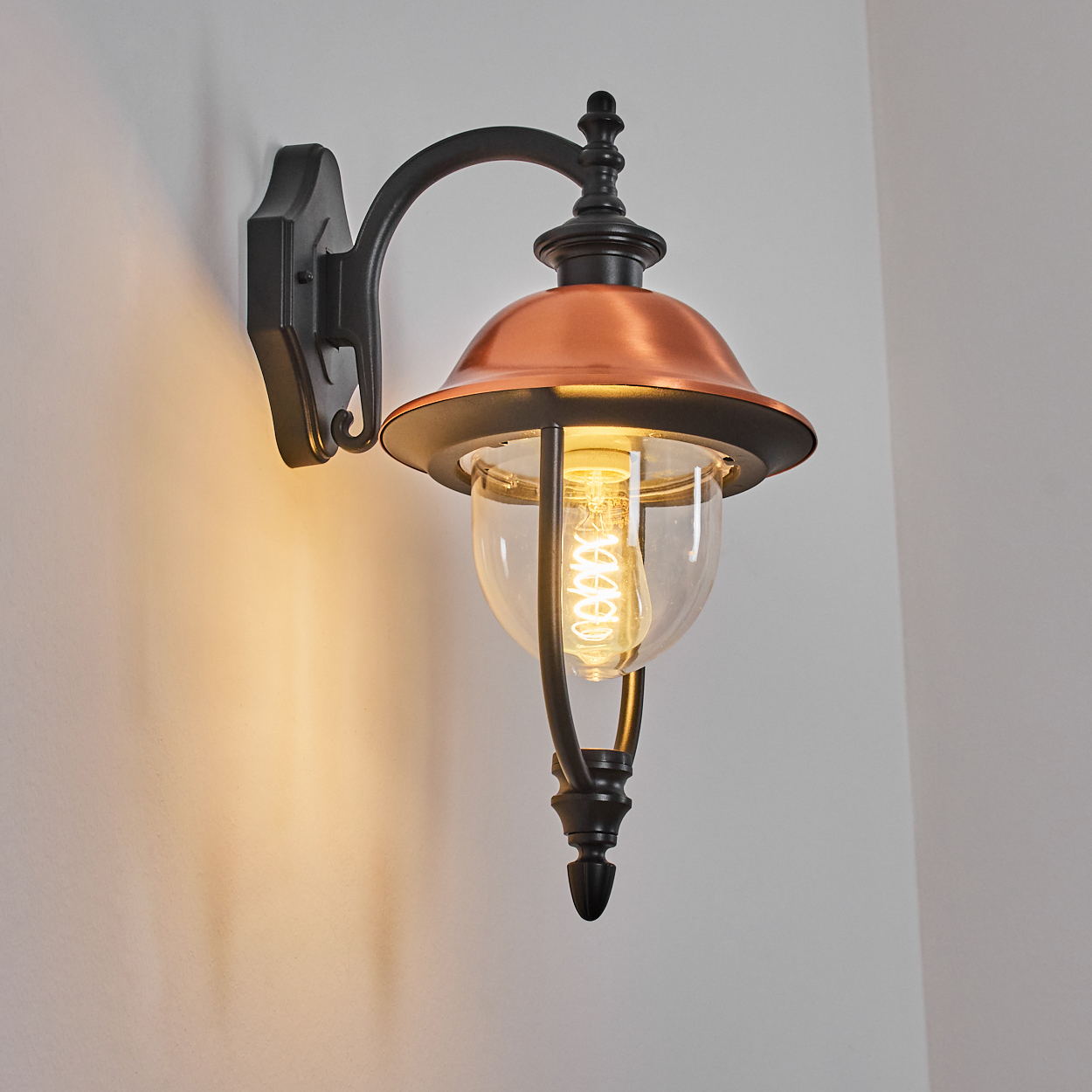 Eine dekorative Wandlampe mit kupferfarbenem Schirm und Glaskugel. Sie ist an einer weißen Wand montiert. Das Licht ist an.