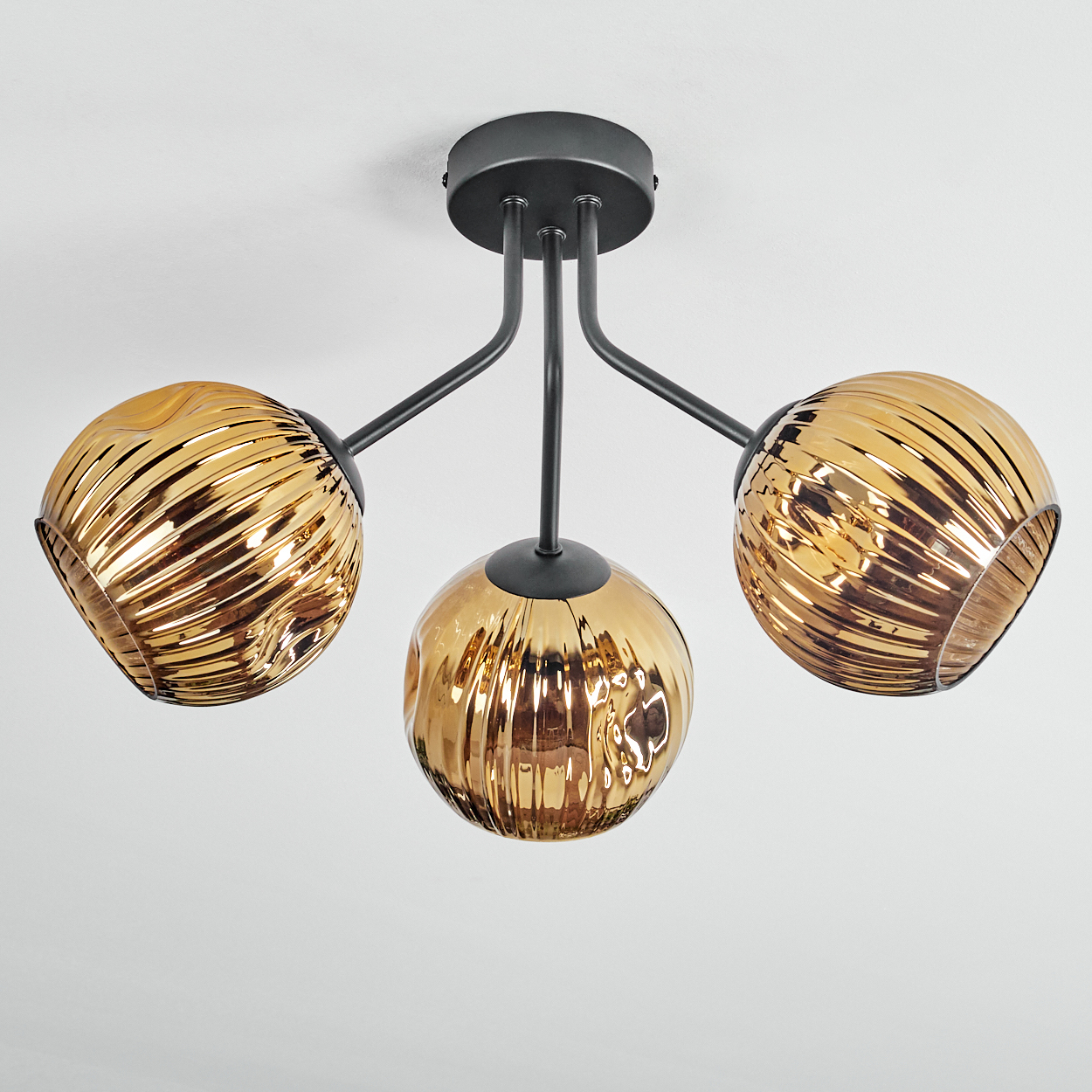 Eine Deckenleuchte mit drei bernsteinfarbenen Glaskugeln. Die Leuchte hat einen schwarzen Metallrahmen und ist an einer weißen Decke befestigt.